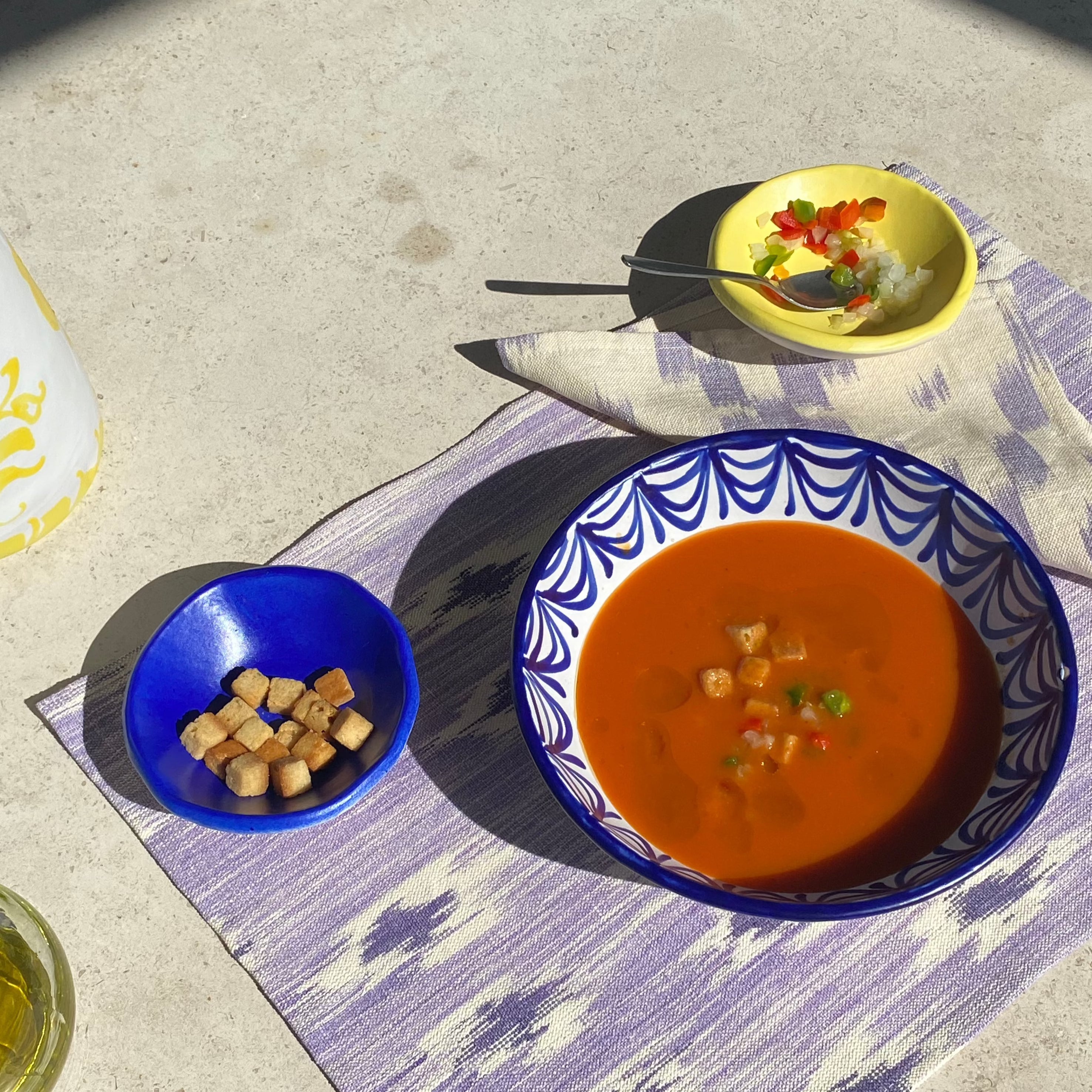 Purple and white handwoven placemat styled with ceramic bowls, gazpacho, croutons and fresh toppings on a sunlit table.