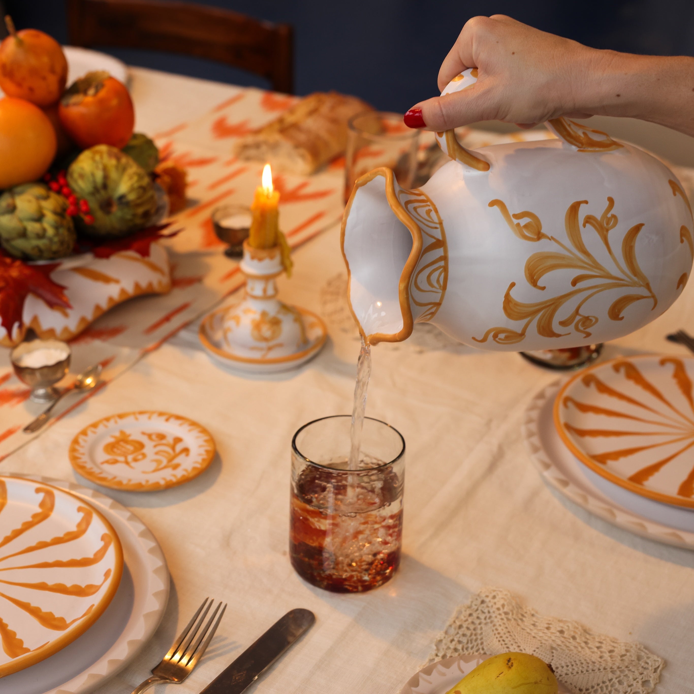 Person pouring liquid from a decorative teapot into a glass on a table set with plates and candles.