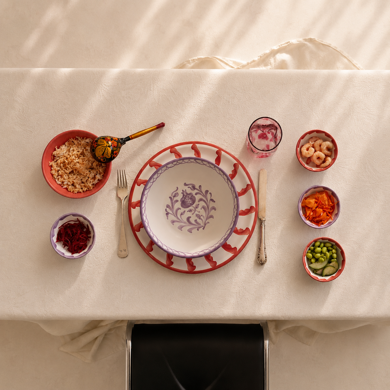 An overhead flat-lay of a dining table set with a central white bowl featuring a purple floral pattern, surrounded by a red and purple decorative plate, smaller matching bowls with food, a glass of pink drink, and silverware on a cream tablecloth. - lifestyle
