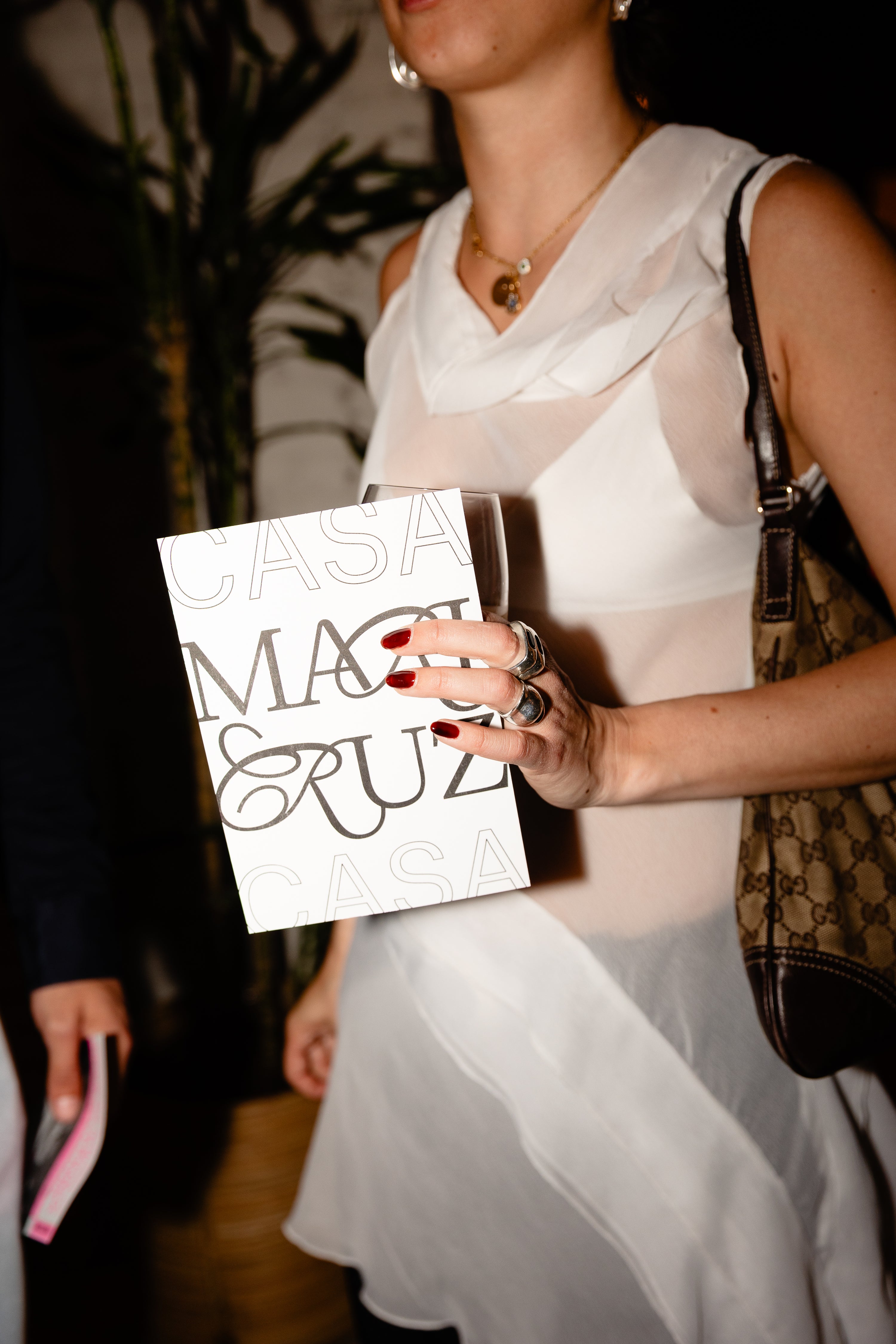 Person holding a white card with text, wearing a white dress and carrying a brown handbag.