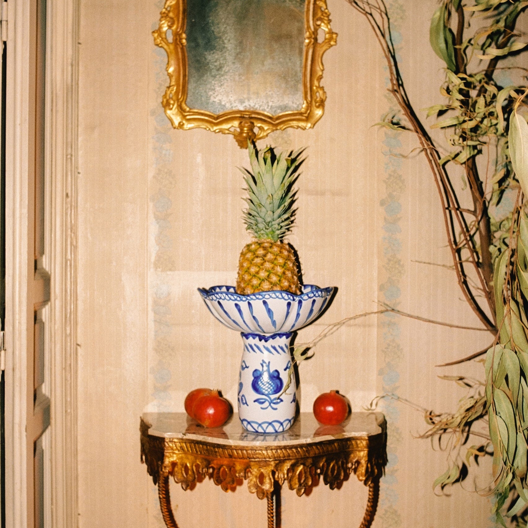 Decorative table with a pineapple on a blue and white granada scallop and leaf shaped bowl, apples, and a gold-framed mirror. - lifestyle