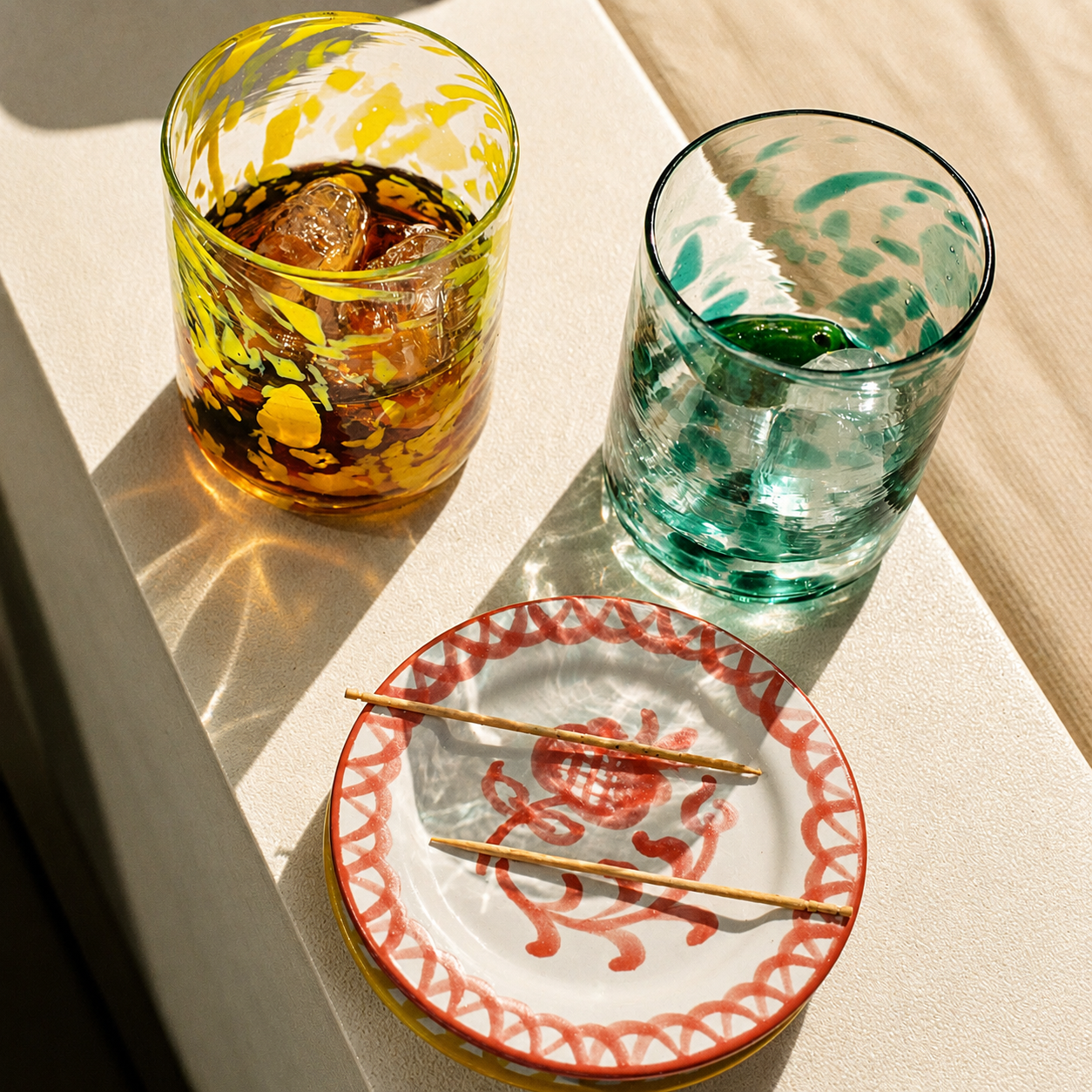 Two patterned drinking glasses and a matching decorative plate arranged on a sunlit tabletop. The left glass contains an amber drink, the right glass is mostly clear with green-blue tinted water, and the plate has red hand-painted motifs with two wooden chopsticks resting on it. -lifestyle