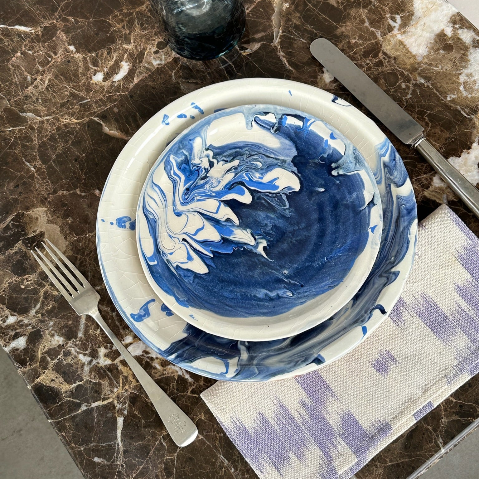 Blue and white hand-painted ceramic plates on a brown marble table, styled with silver cutlery, a glass and a lilac patterned cocktail napkin. - lifestyle