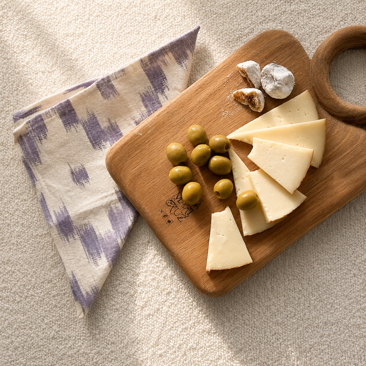 A wooden serving board with a handle holds wedges of cheese, green olives, and a few pieces of white chocolate, styled on a textured cream fabric next to a purple and cream patterned cloth napkin. - lifestyle