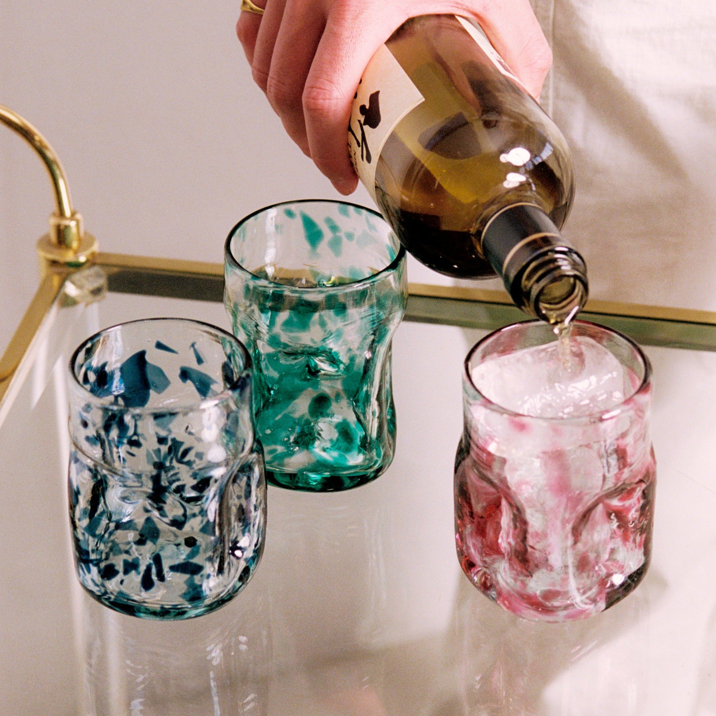 Person pouring a drink into a glass with ice, surrounded by speckled glasses on a reflective surface. - lifestyle