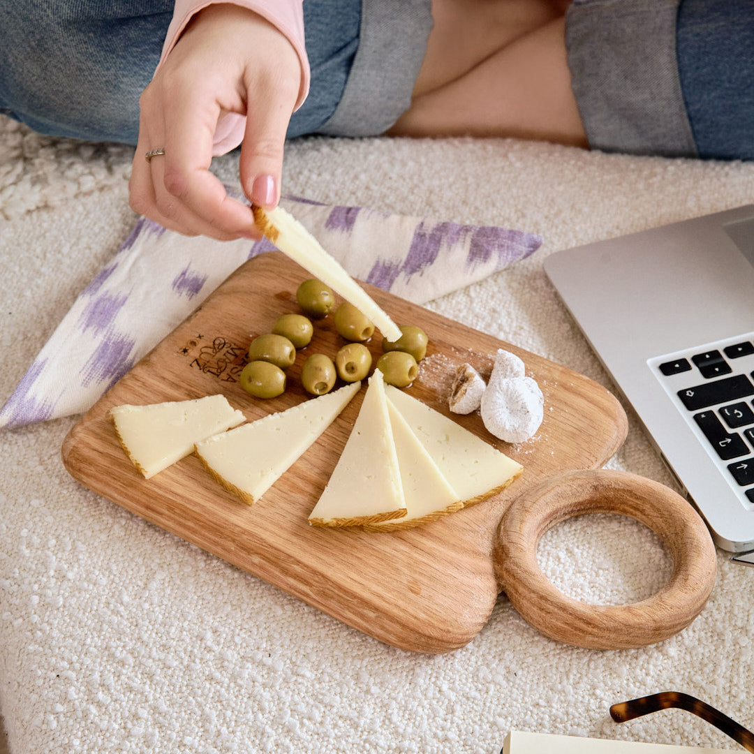Person eating cheese and olives on a wooden board with a laptop and glasses in the background.