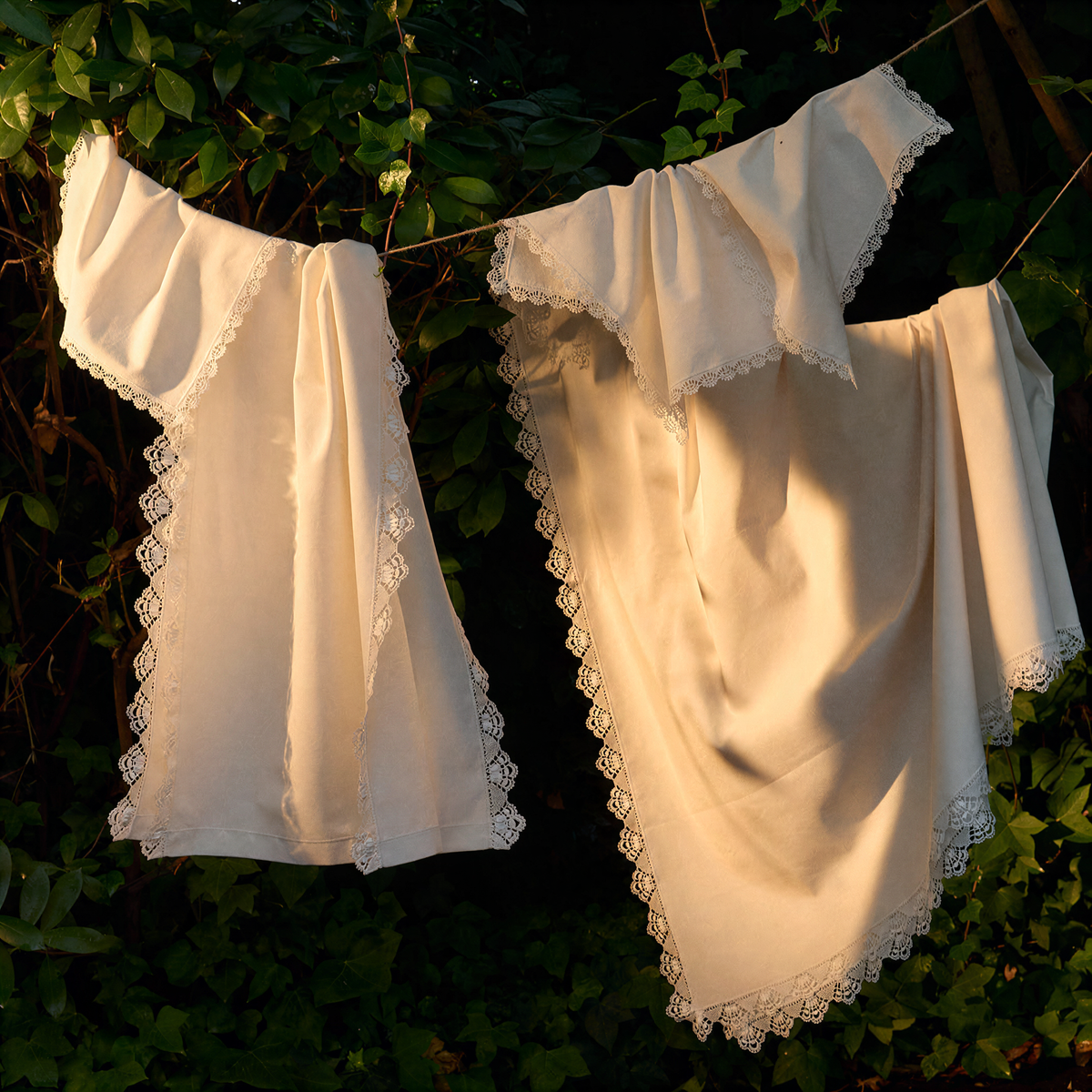 Two cream-colored cotton napkins with delicate lace trim hanging from a string outdoors against a backdrop of dark green foliage. - lifestyle