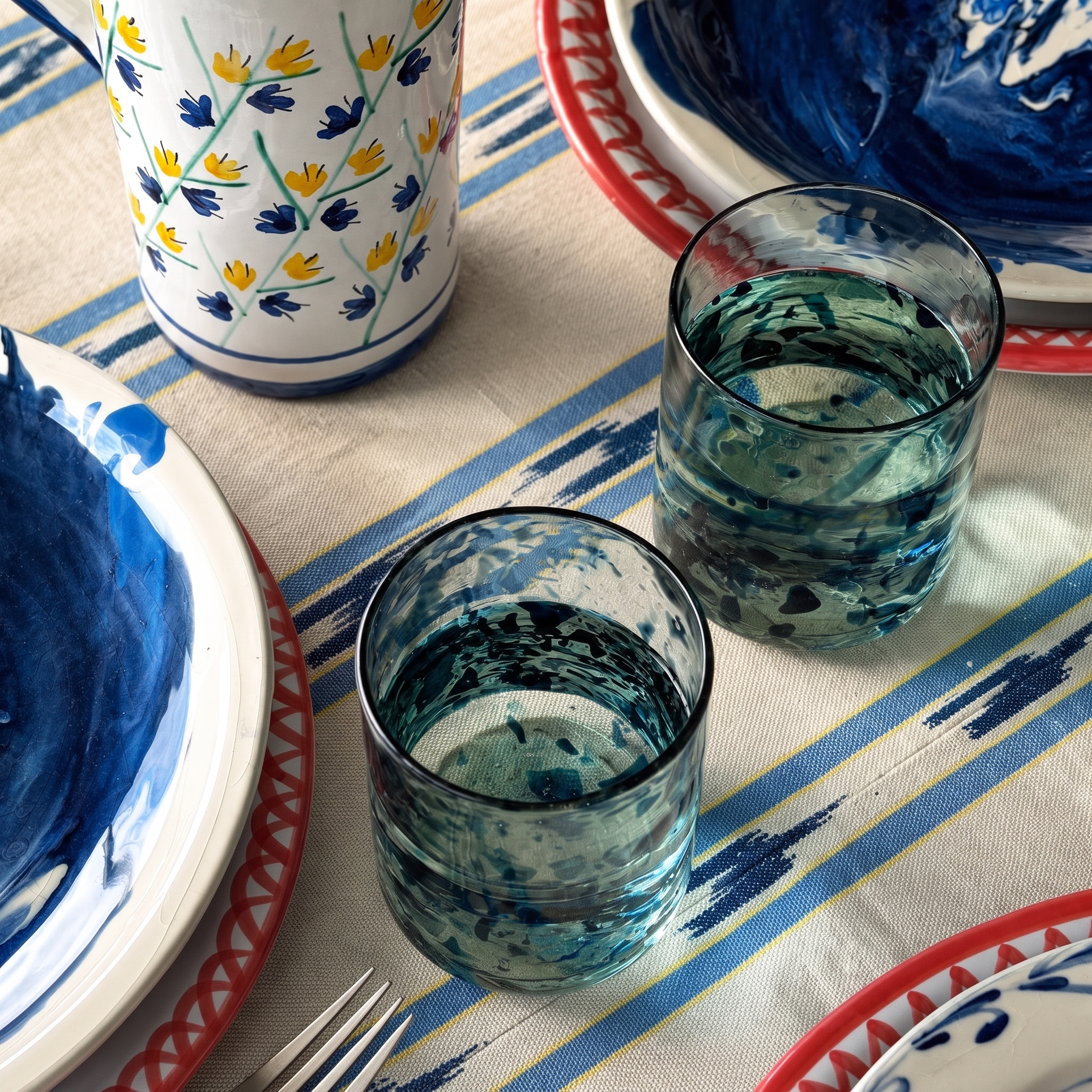 Two textured blue and white marbled glass tumblers filled with water, set on a striped tablecloth alongside matching blue and white patterned ceramic plates and a floral vase. - lifestyle