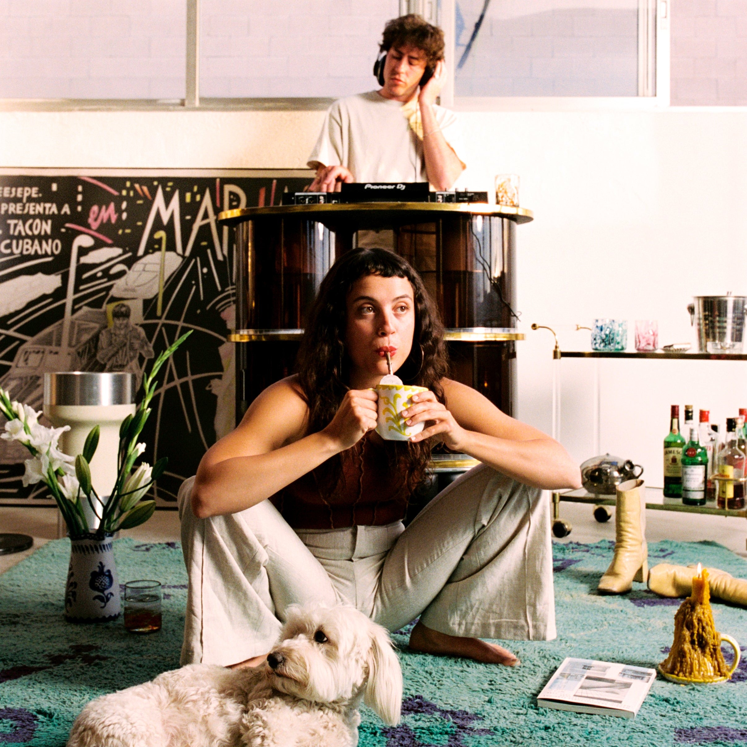 Person sitting on a colorful rug with a dog, drinking from a cup, in a room with a person playing music on a turntable. - lifestyle
