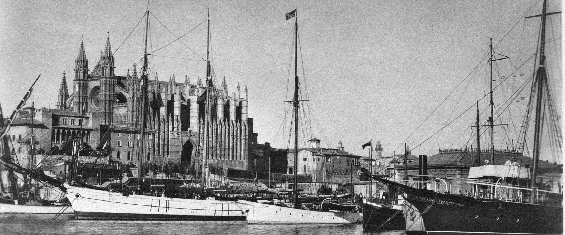 Historical black and white photograph of a harbor with ships and a large building, possibly a cathedral.