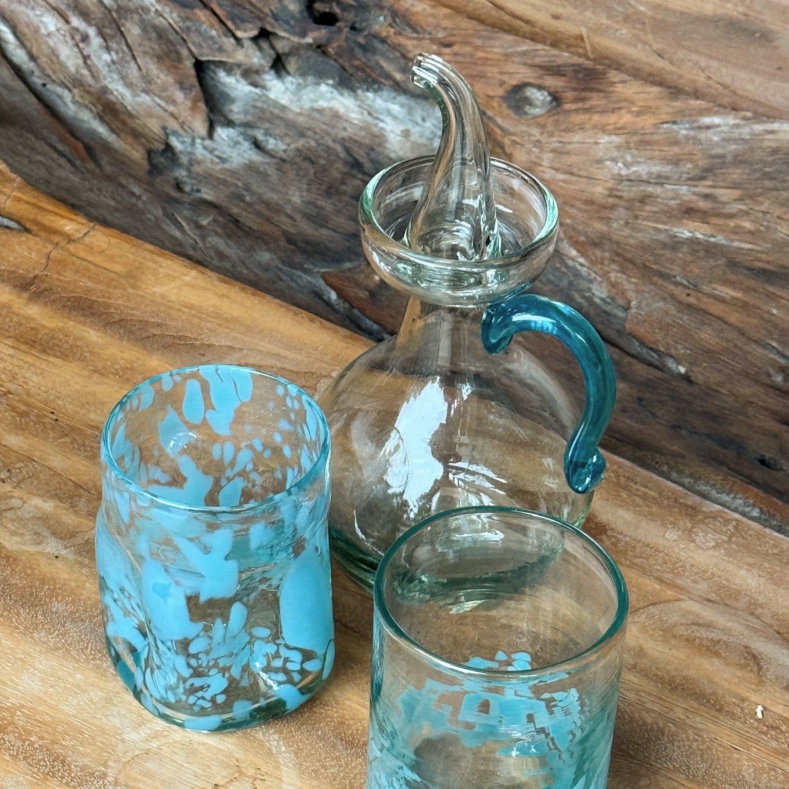 Two turquoise glass tumblers and a clear glass carafe with a blue handle on a wooden surface.