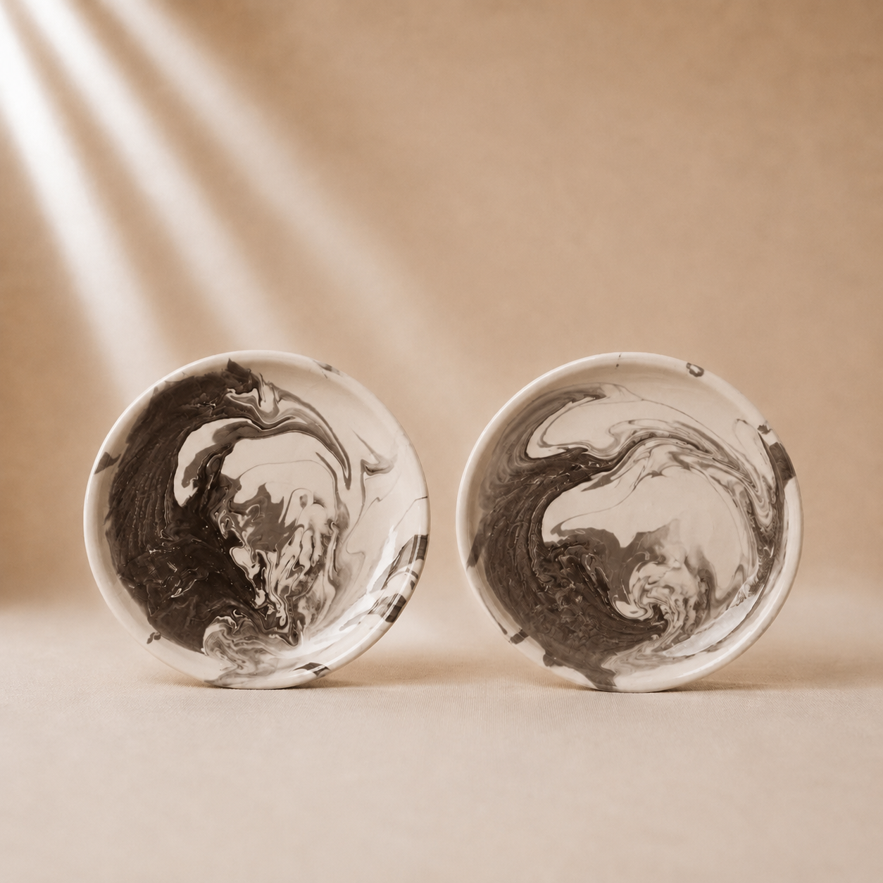 Two small round white bowls with black and gray marbled swirl patterns, displayed side-by-side on a beige surface with soft light rays in the background.