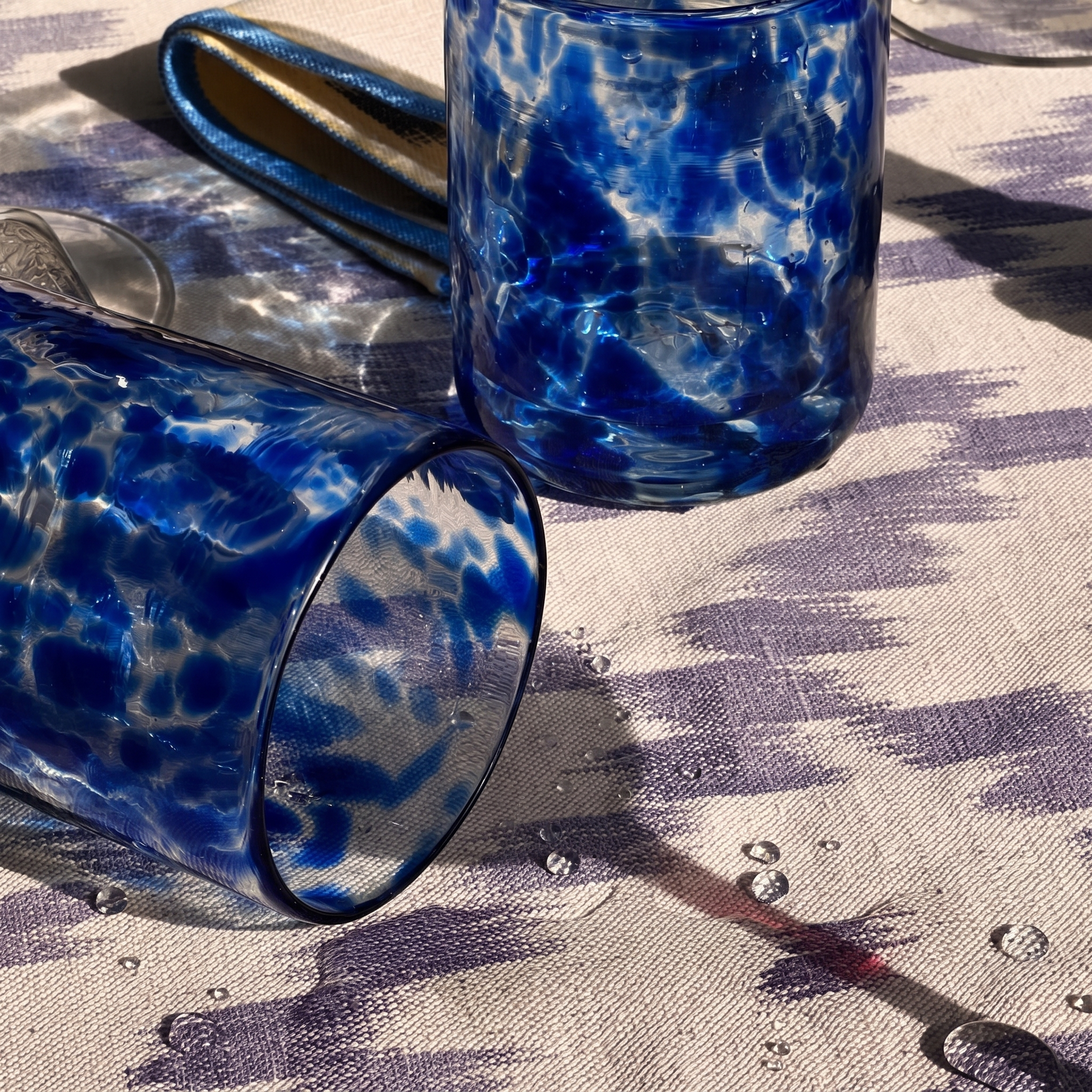 Two blue and white marbled glass tumblers resting on a patterned tablecloth with water droplets, alongside a metallic blue napkin. - lifestyle