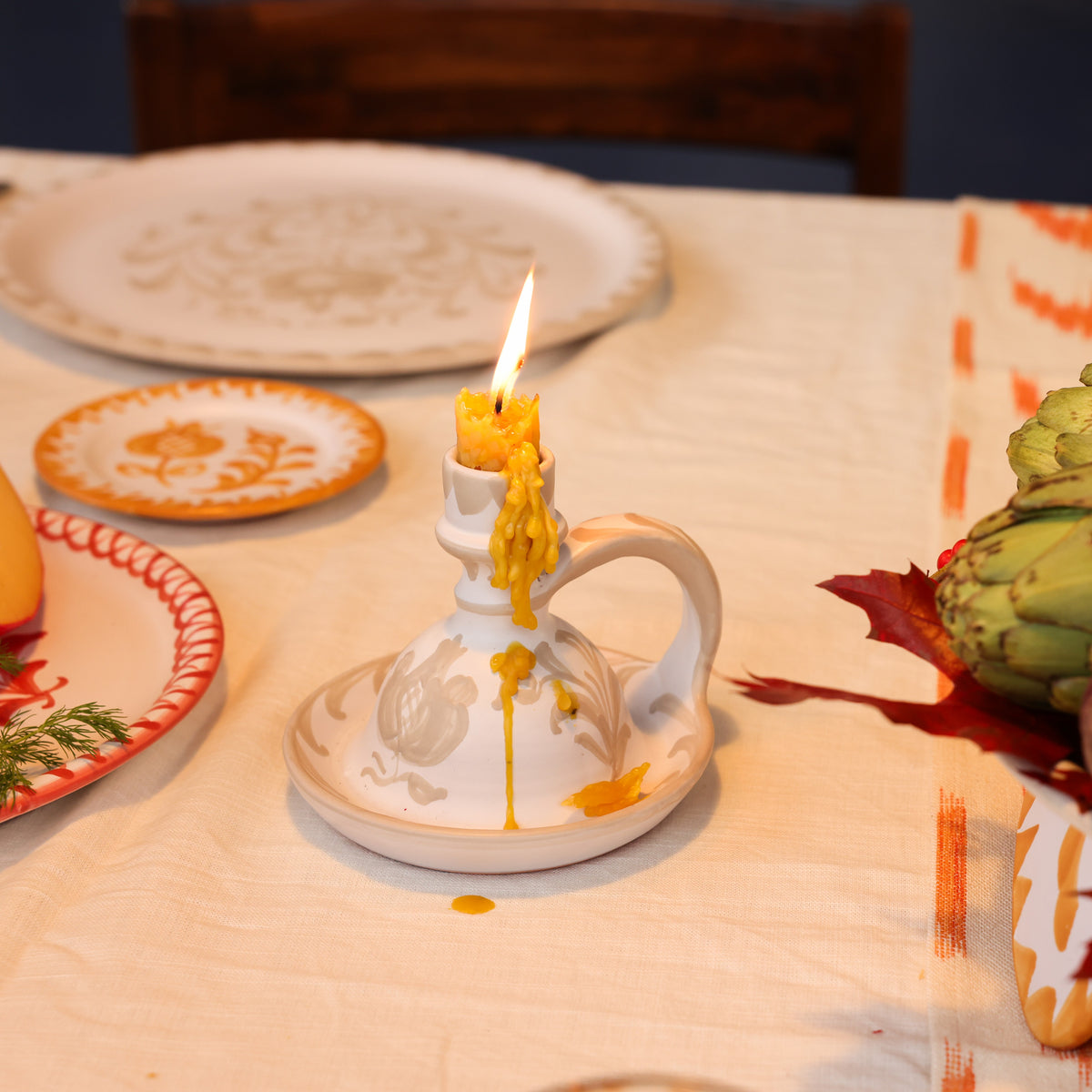 A white ceramic candle holder with a handle and matching saucer, holding a lit yellow candle with melted wax dripping down. The holder is set on a cream tablecloth with autumn-themed tableware, a corn cob, and an artichoke in the background. - lifestyle