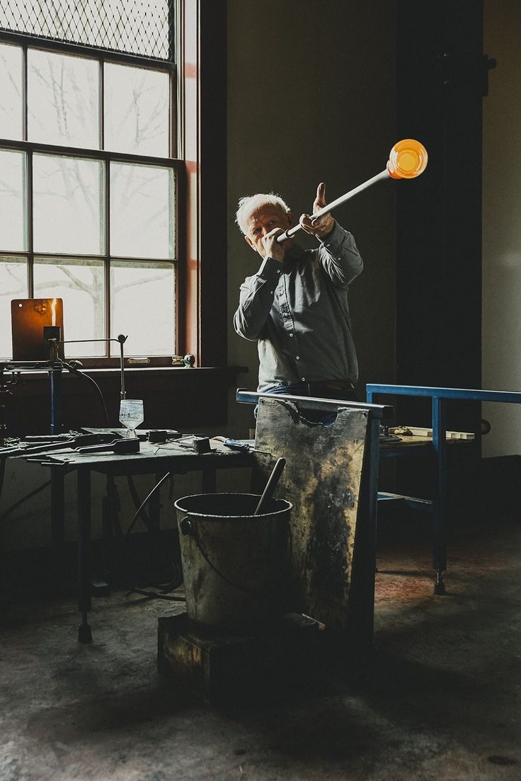 Glassblower shaping molten glass in a workshop with large windows.