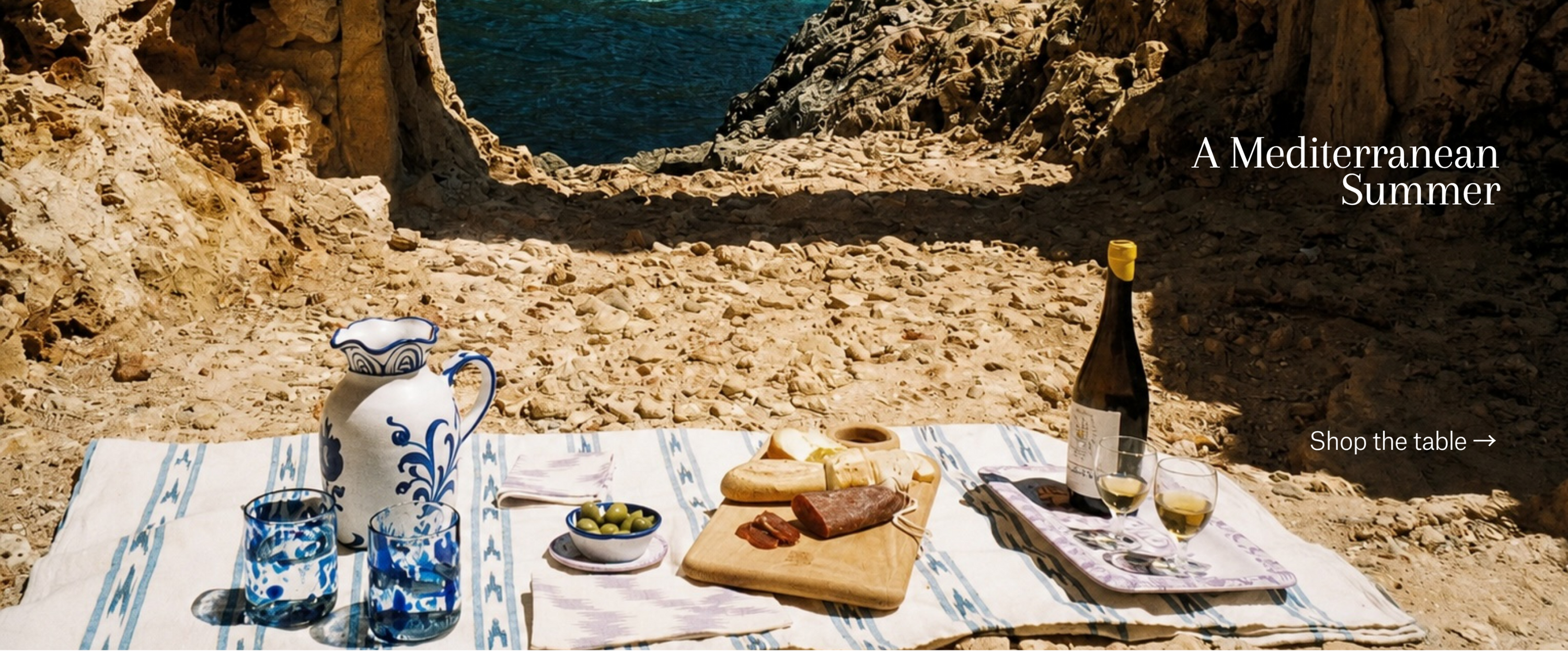 Table setting with food and drinks by a rocky coastal area, featuring a bottle of wine and a pitcher.