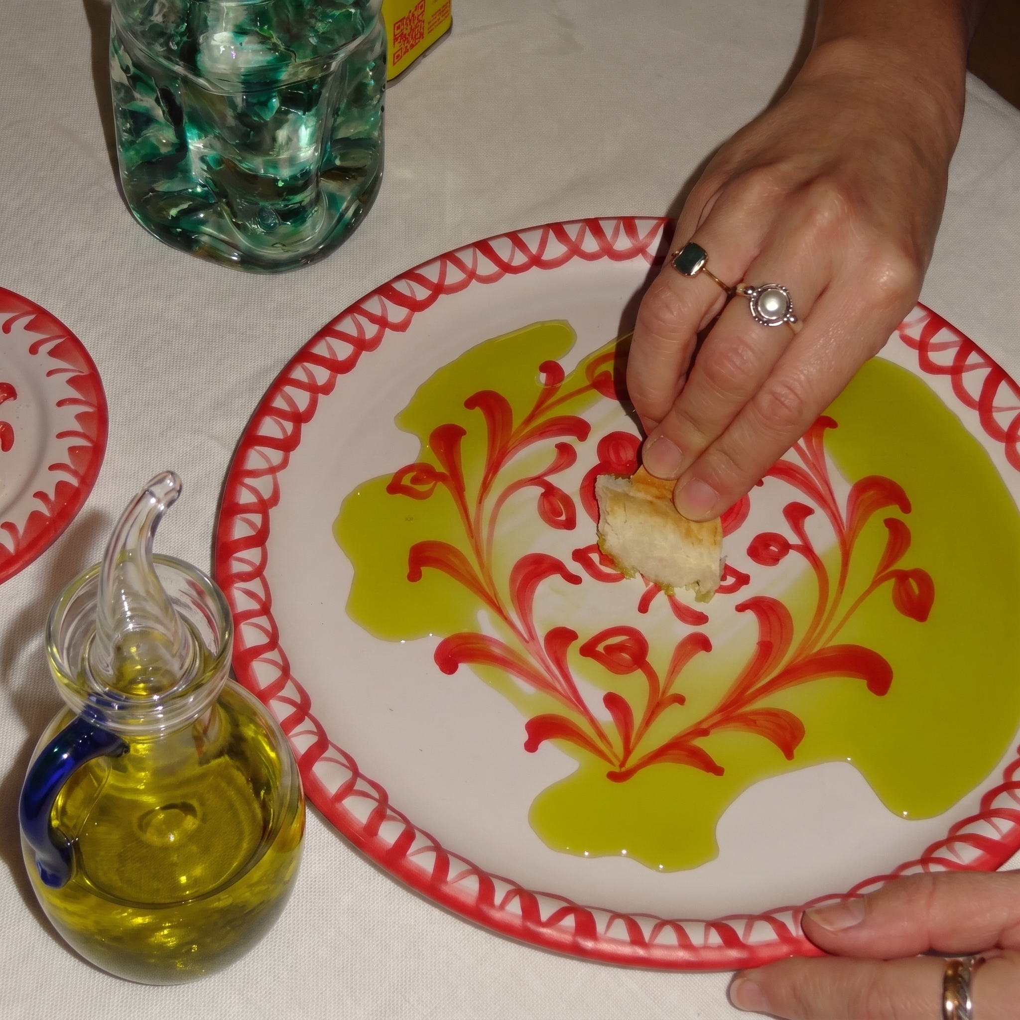 A close-up of a hand dipping a piece of bread into olive oil on a white ceramic plate with a red patterned rim, with a small glass oil bottle nearby on a white tablecloth. - lifestyle