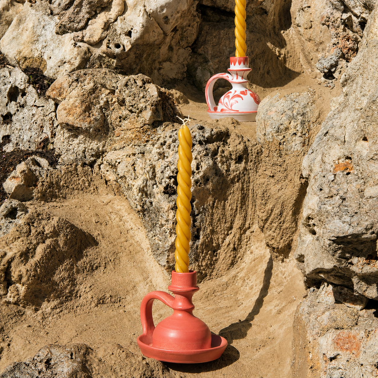 Two decorative ceramic candle holders shaped like small red-orange pitchers, each holding a twisted yellow candle, set against a rocky sandy outdoor background. - lifestyle