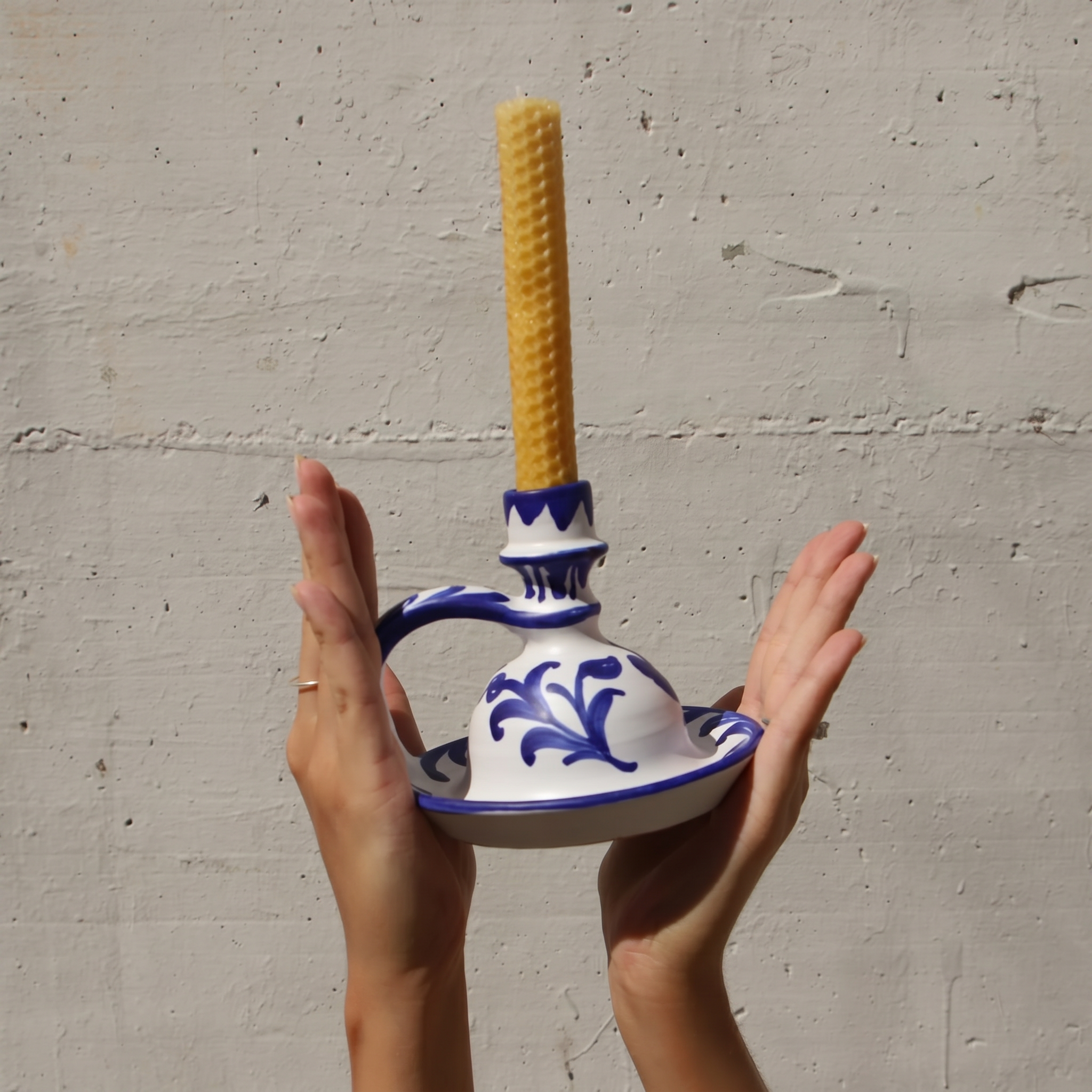 A person holds a white ceramic candle holder with blue floral patterns and a yellow textured candle inserted, set against a light gray concrete wall. - lifestyle