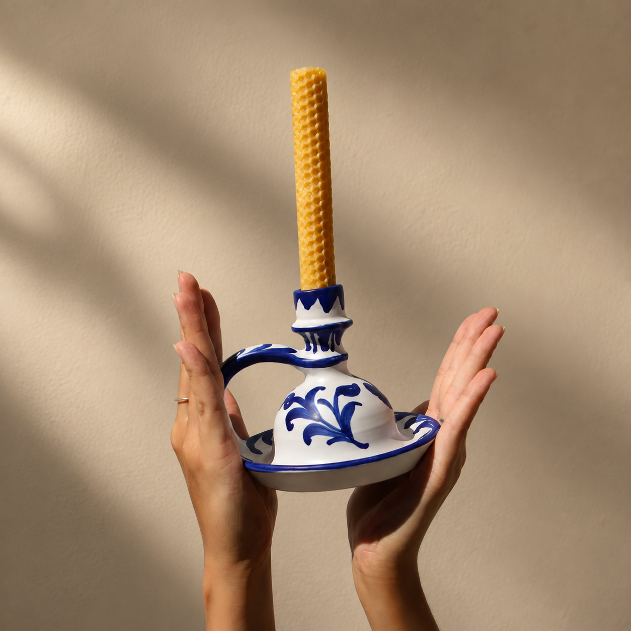 A person holds a white ceramic candle holder with blue floral patterns and a tall yellow candle, set against a warm beige background with soft leaf shadows. - lifestyle
