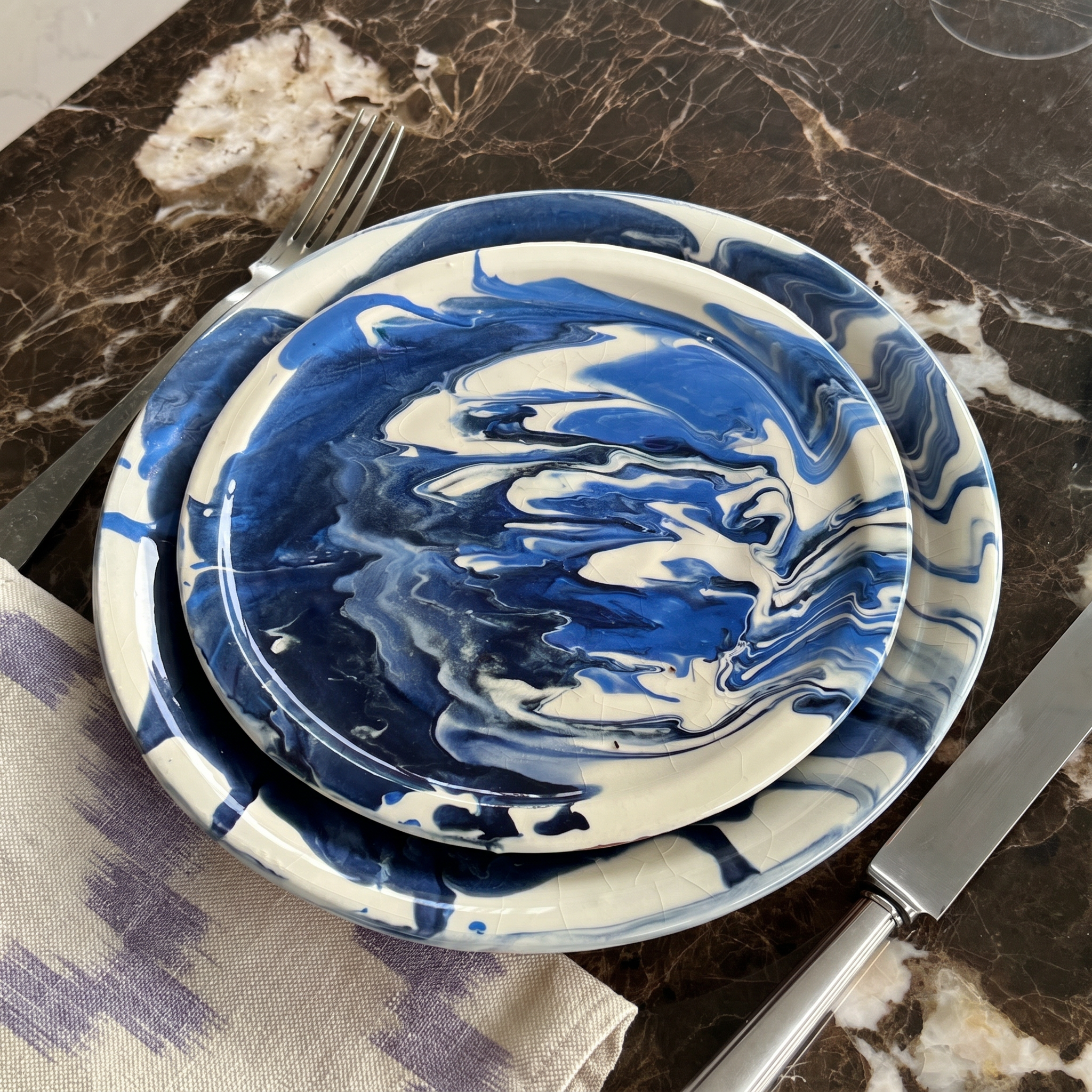 A stack of two round plates with a blue and white marbled swirl pattern, styled on a dark marble tabletop with a fork and knife. - lifestyle