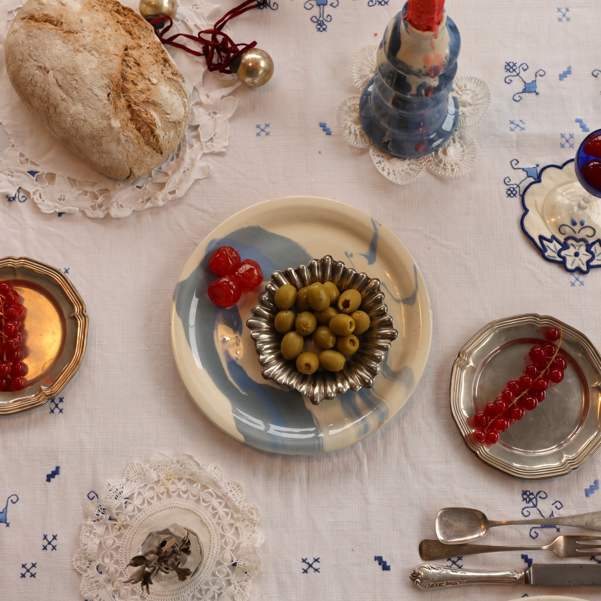 A styled tabletop scene on a white embroidered tablecloth with a rustic loaf of bread, a blue-and-white ceramic bell, a small plate of green olives in a silver bowl, red berries on silver plates, and silver cutlery. - lifestyle