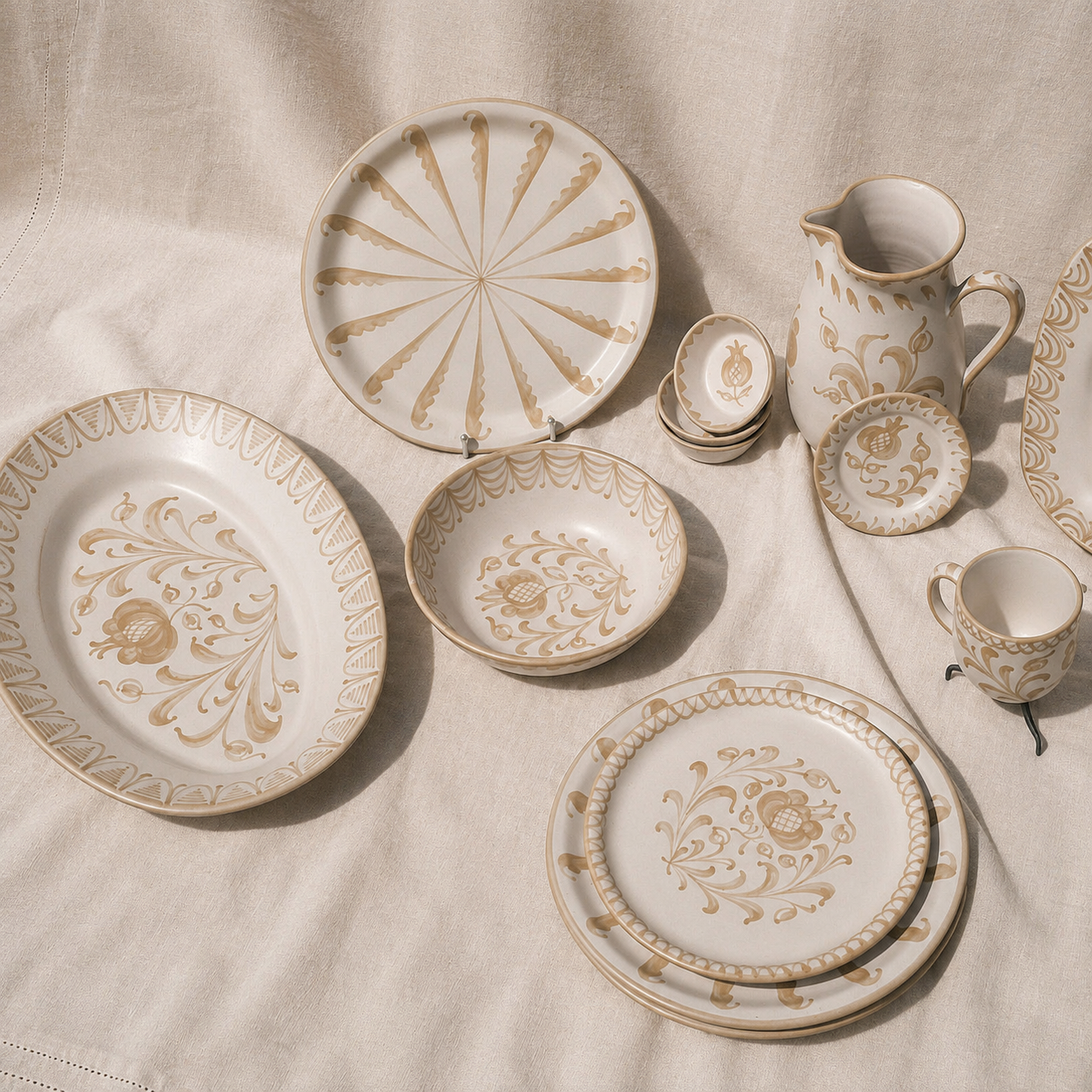 A coordinated ceramic tableware set arranged on a cream fabric surface, including plates, bowls, a pitcher, and cups with a warm beige ornamental floral and scroll pattern. - lifestyle