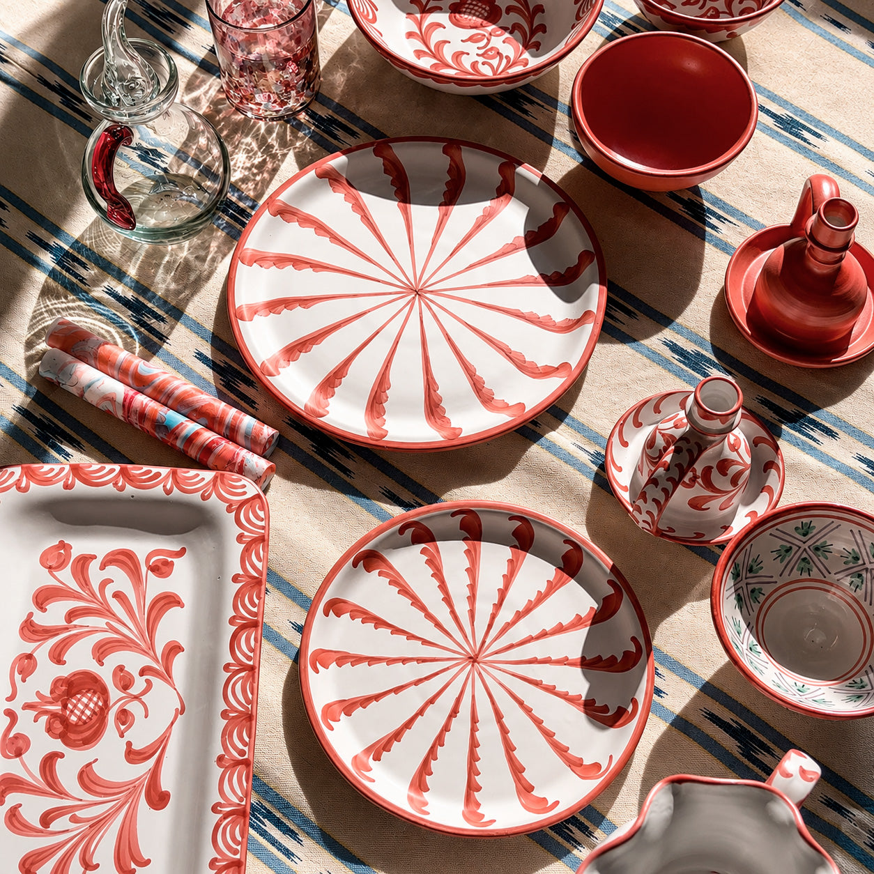 A coordinated ceramic tableware set arranged on a striped textile. The set includes round and rectangular plates, bowls, a small vase, and a serving tray, all in white with red decorative patterns and some gray interior detailing.