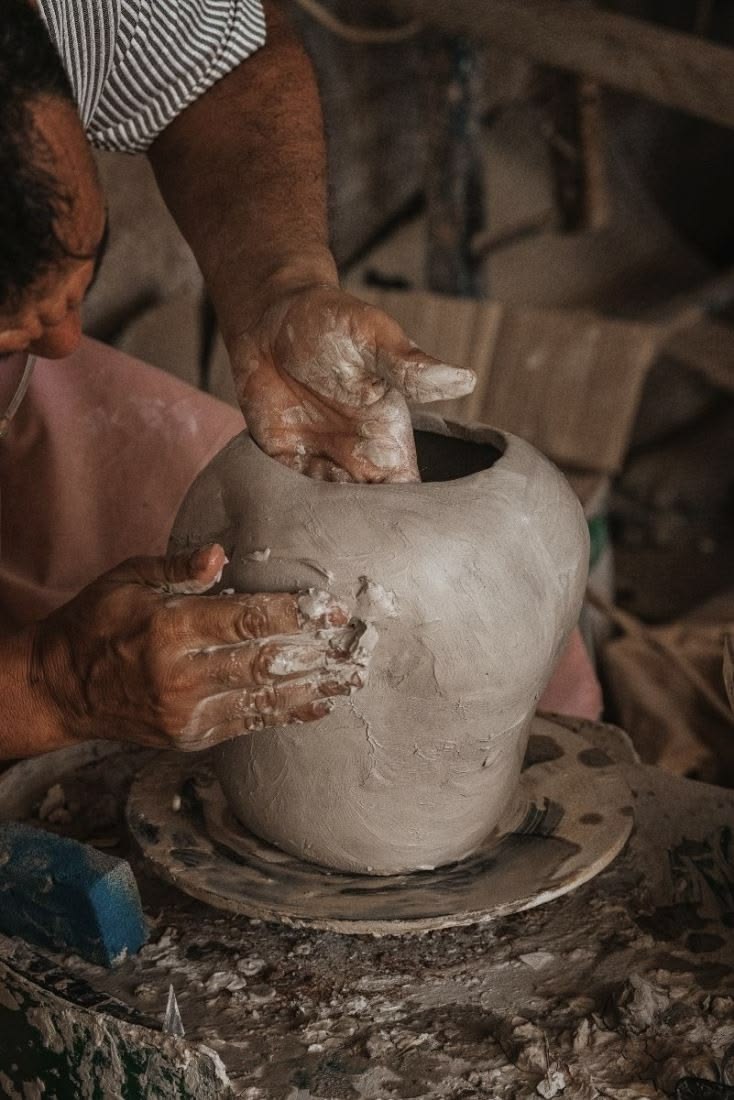 Person shaping a clay pot on a pottery wheel in a workshop setting.