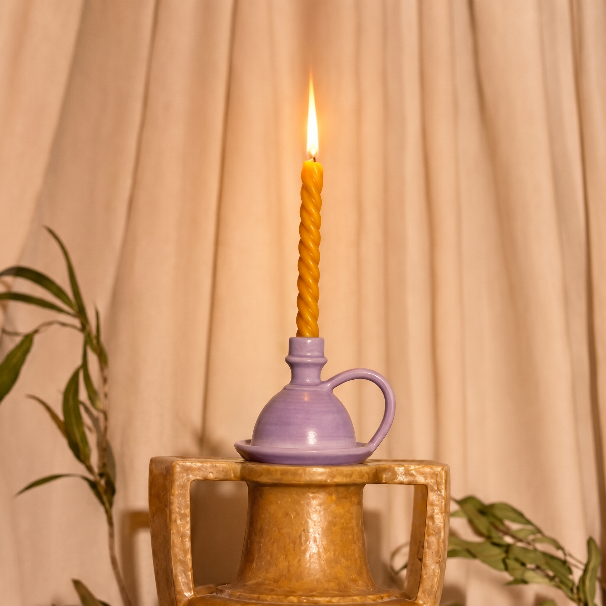 A lit twisted yellow candle in a small lavender ceramic holder, displayed on a rustic gold-toned pedestal against a draped beige fabric backdrop with soft plant leaves at the sides. - lifestyle