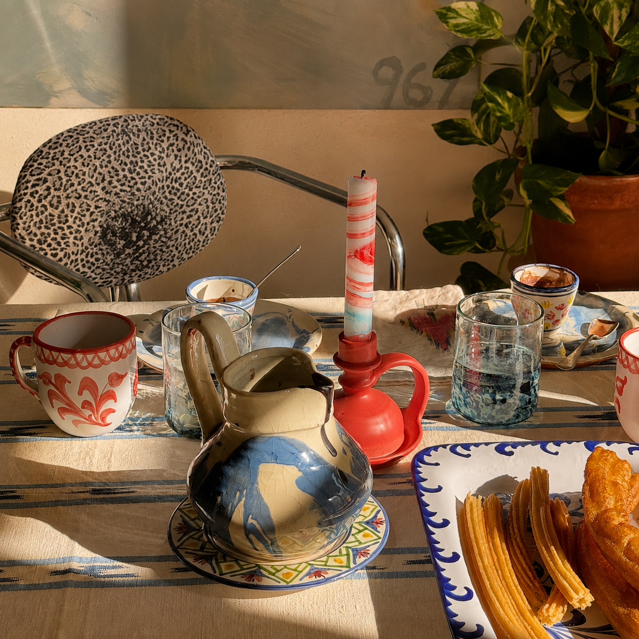 A styled tabletop scene with ceramic tableware, including a blue and beige pitcher, patterned mugs, a red candle holder, and a plate of churritos, set in a warm indoor environment with sunlight, a plant, and a patterned chair visible in the background. - lifestyle