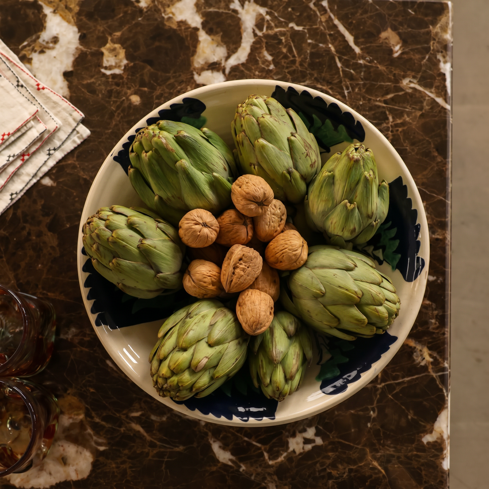 A white ceramic bowl with a blue scalloped rim, filled with green artichokes and topped with whole walnuts, set on a dark brown marble surface. - lifestyle