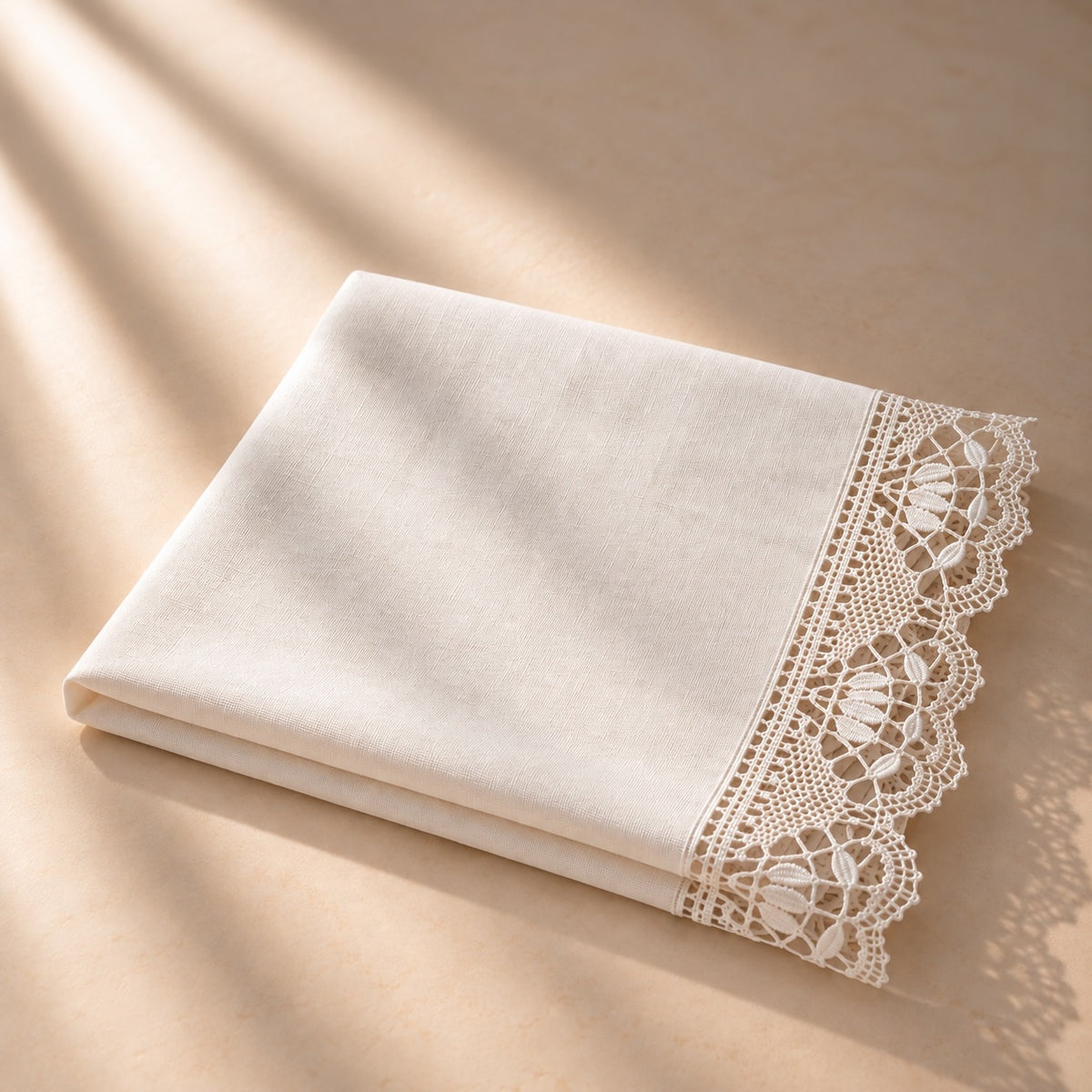 A folded cream-colored fabric item, likely a pillowcase or napkin, featuring a decorative white lace border, displayed on a warm beige surface with soft diagonal shadows.
