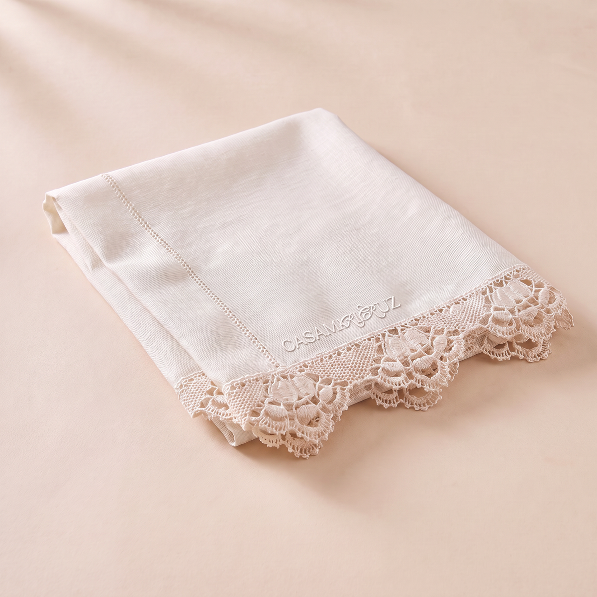 An off-white folded cotton handkerchief with a delicate floral lace trim and a subtle embroidered brand name, displayed on a soft beige background.