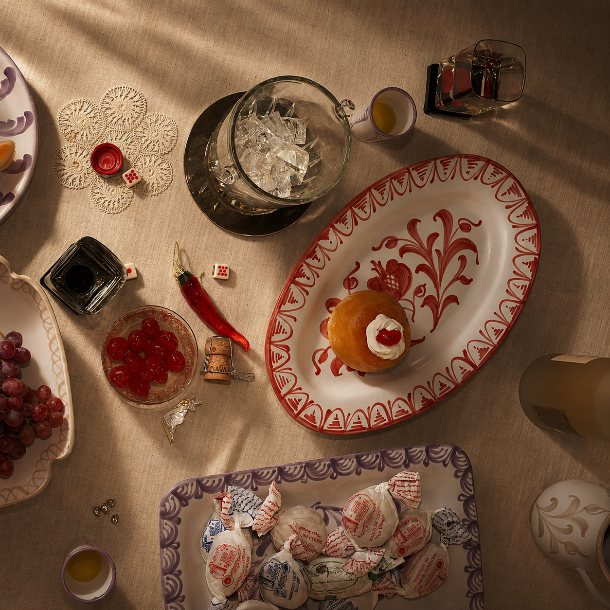 An overhead styled tabletop scene on a beige tablecloth with multiple decorative dishes, a glass pitcher of iced drink, candies, grapes, a red chili pepper, dice, and small cups. The central salable subject appears to be the large oval white plate with red floral scrollwork and a red decorative border, holding a small pastry topped with cream and a red heart. - lifestyle