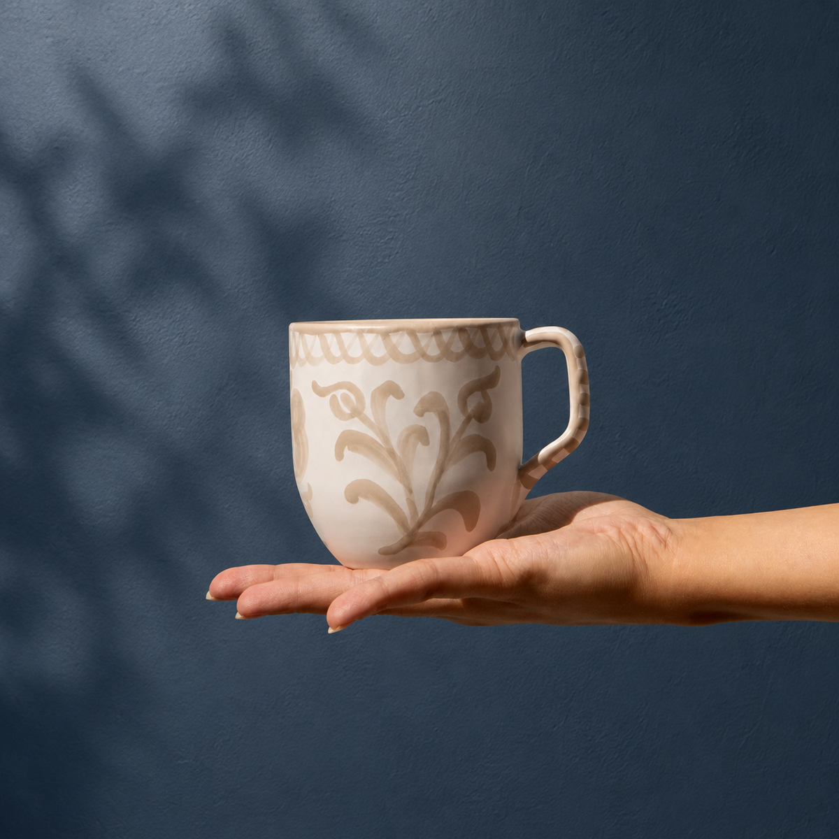 A hand holding a ceramic mug with a beige floral pattern on a warm off-white background, set against a dark blue textured wall with soft leaf shadows. - lifestyle
