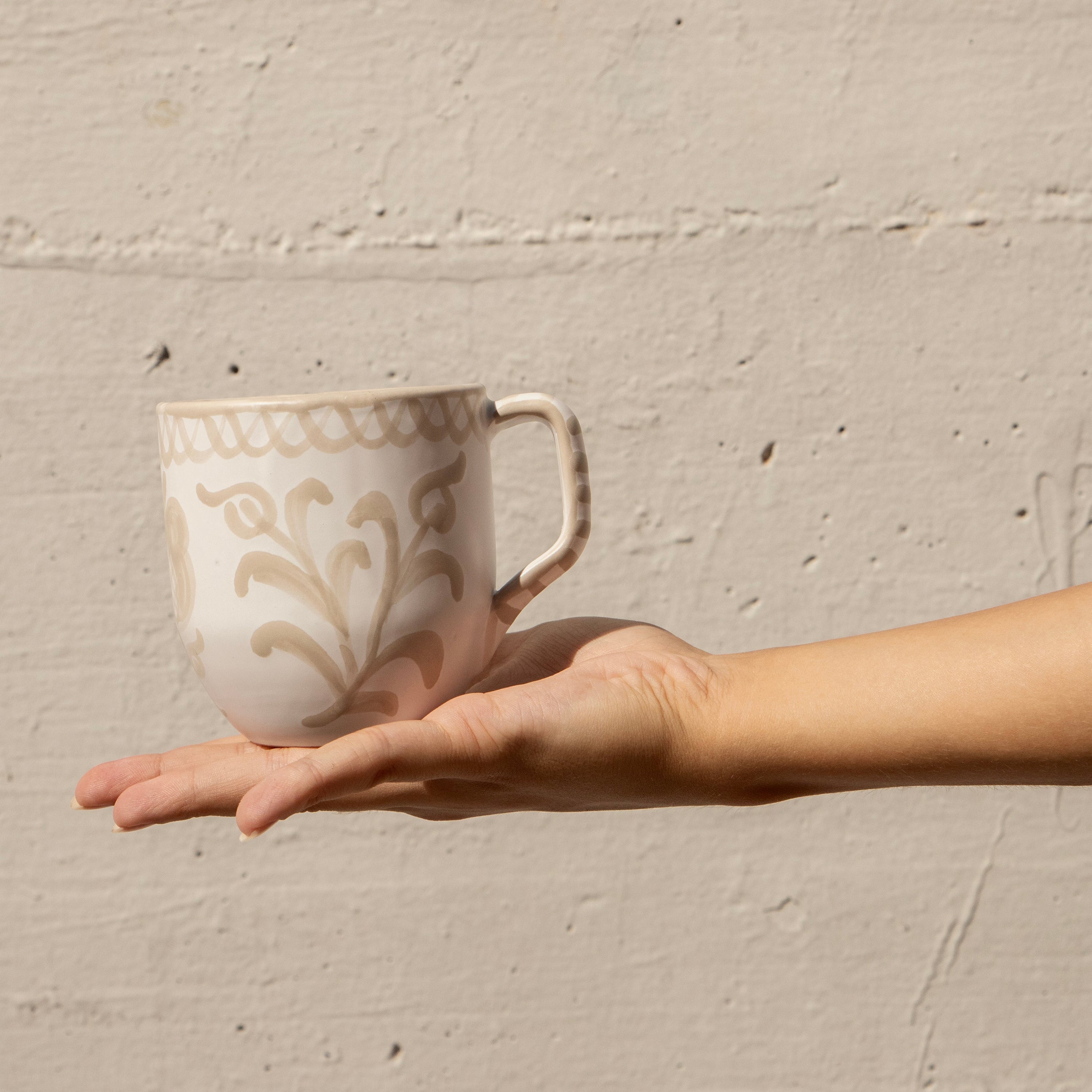 Hand holding a ceramic mug with decorative patterns against a beige wall.