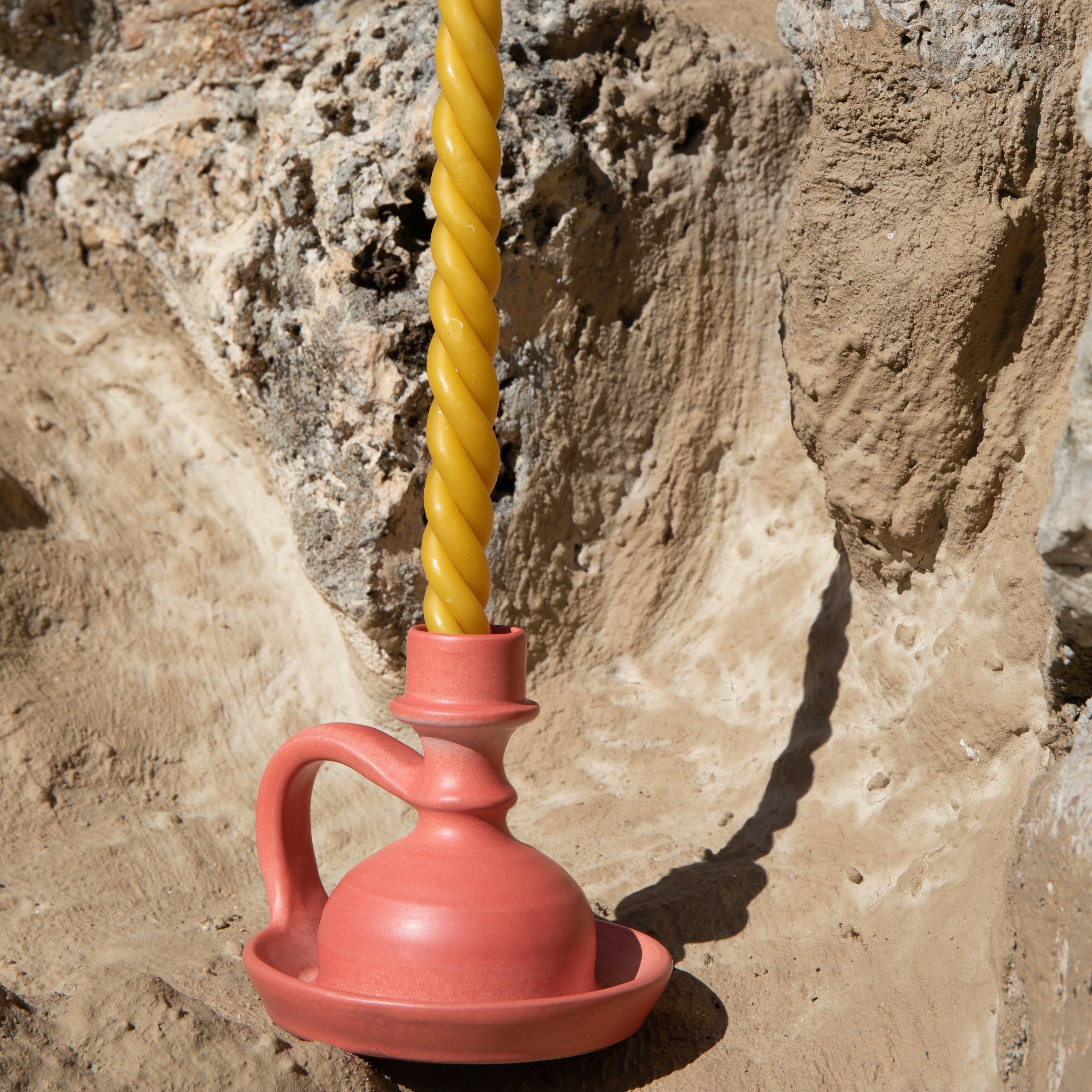 Yellow twisted candle in a red ceramic holder against a stone wall.