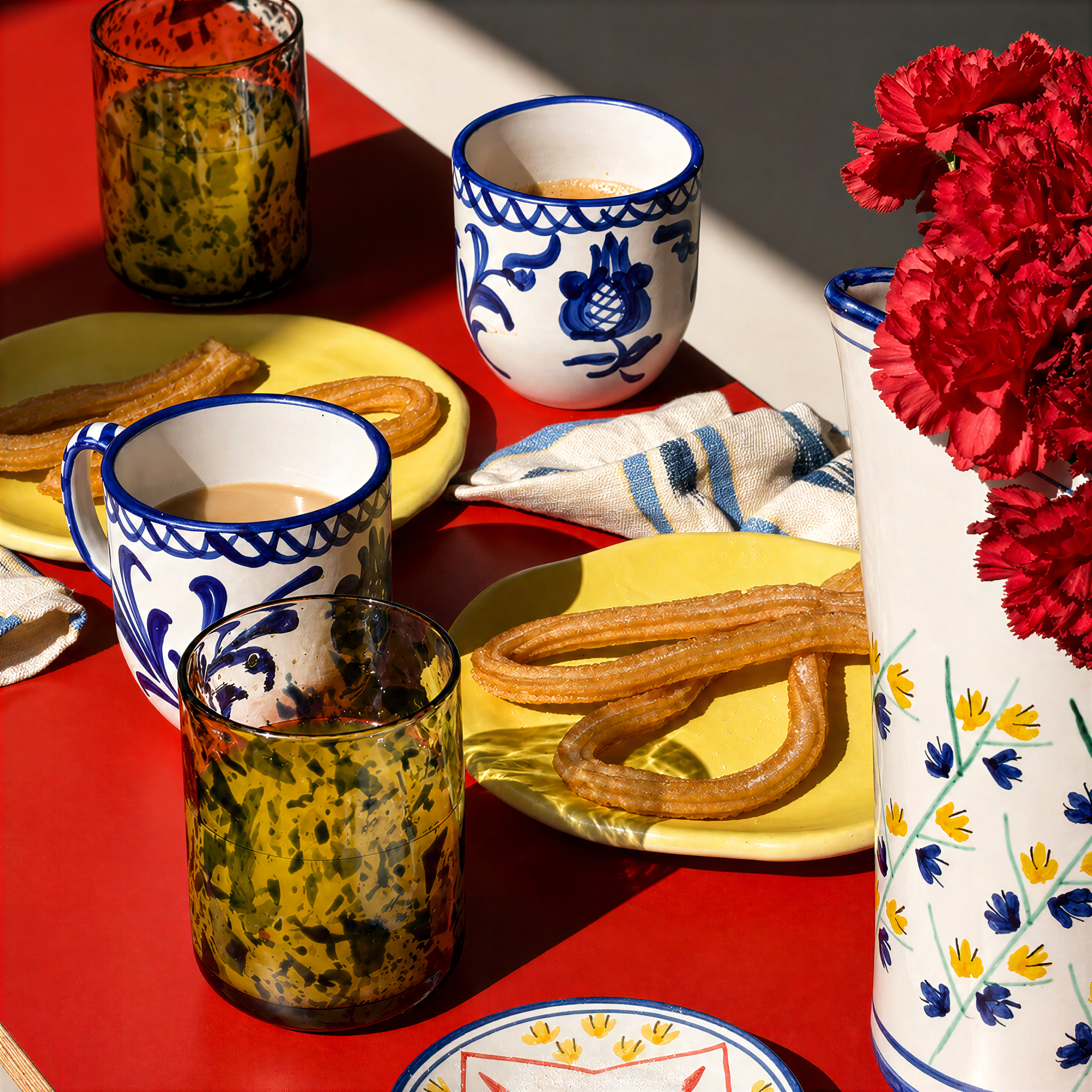 A styled tabletop scene on a white embroidered tablecloth with churros, a blue-and-white ceramic bell, a small plate of green olives in a silver bowl, red berries on silver plates, and silver cutlery. - lifestyle
