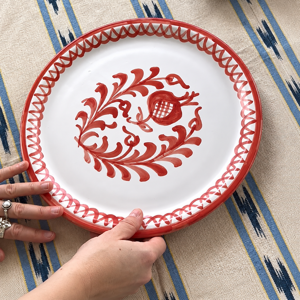 A round white ceramic serving platter with a red painted floral scroll pattern and a red rim, held by two hands over a blue and beige patterned textile. - lifestyle