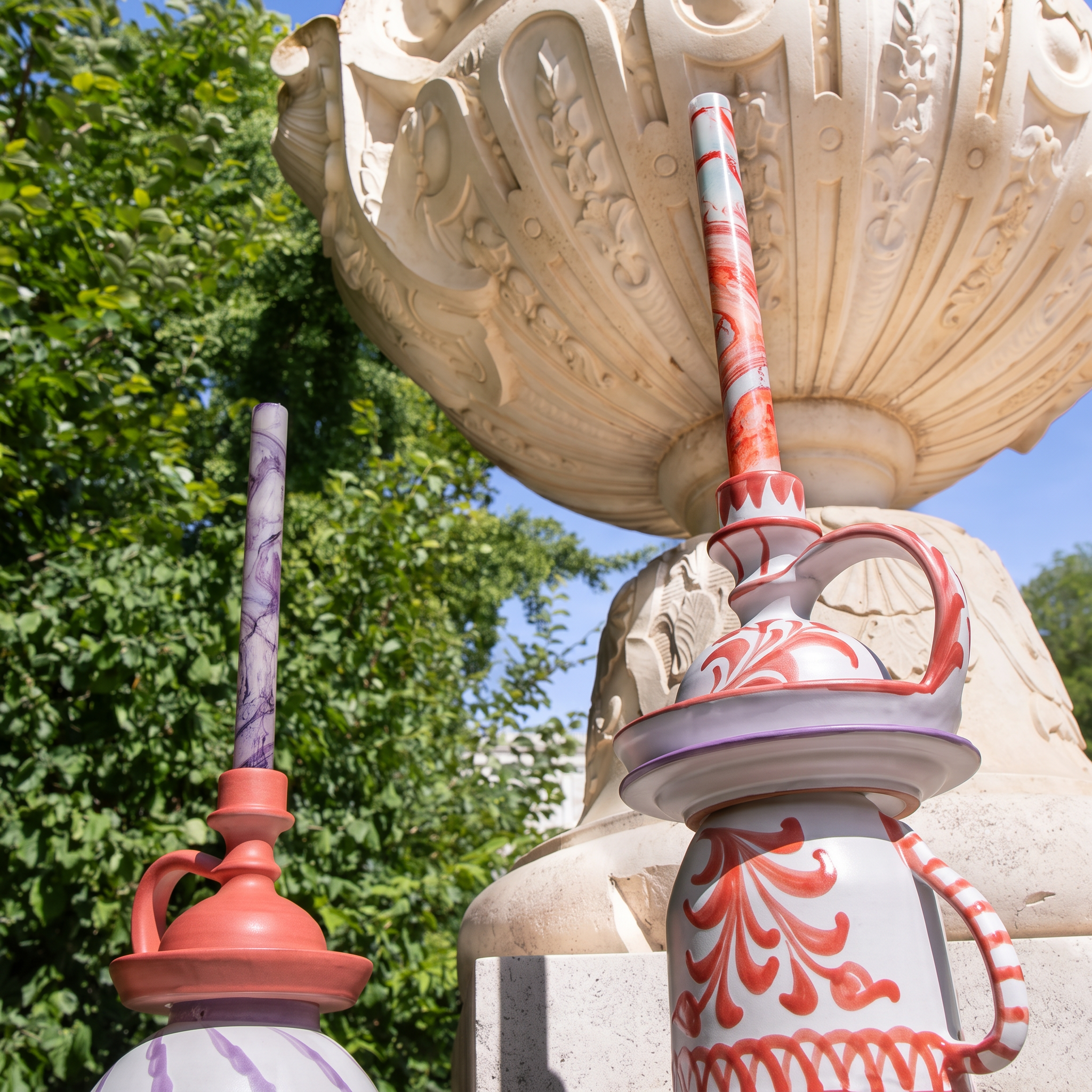 Decorative ceramic drinking glasses with red scrollwork and purple striped handles, styled outdoors in front of a large stone fountain structure and green trees. - lifestyle