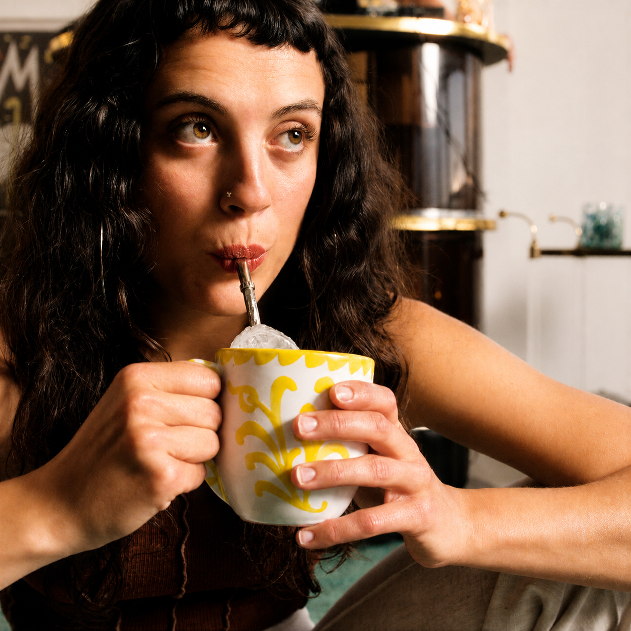 A woman with dark curly hair holds a white ceramic mug with a yellow floral pattern while drinking from a metal spoon in a cup of hot chocolate or cocoa inside a cozy indoor setting. - lifestyle
