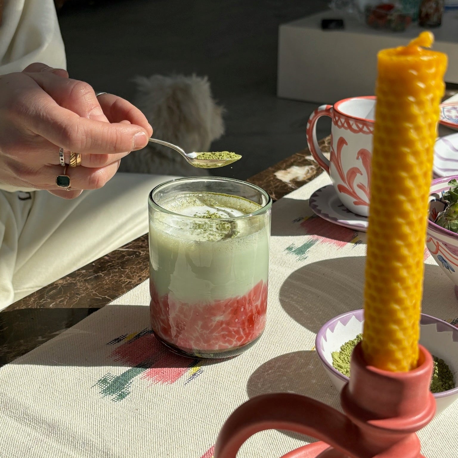 Person spooning a green substance into a glass on a table with a pink teapot and yellow candle. - lifestyle