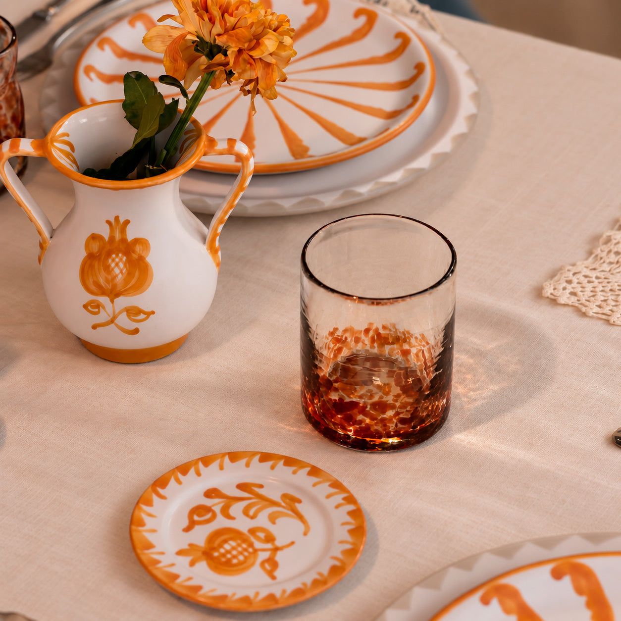 A styled tabletop scene with white ceramic tableware decorated in orange, including a small vase with orange flowers, stacked plates, a small round plate, and a textured clear tumbler with an orange patterned base on a beige tablecloth. - lifestyle