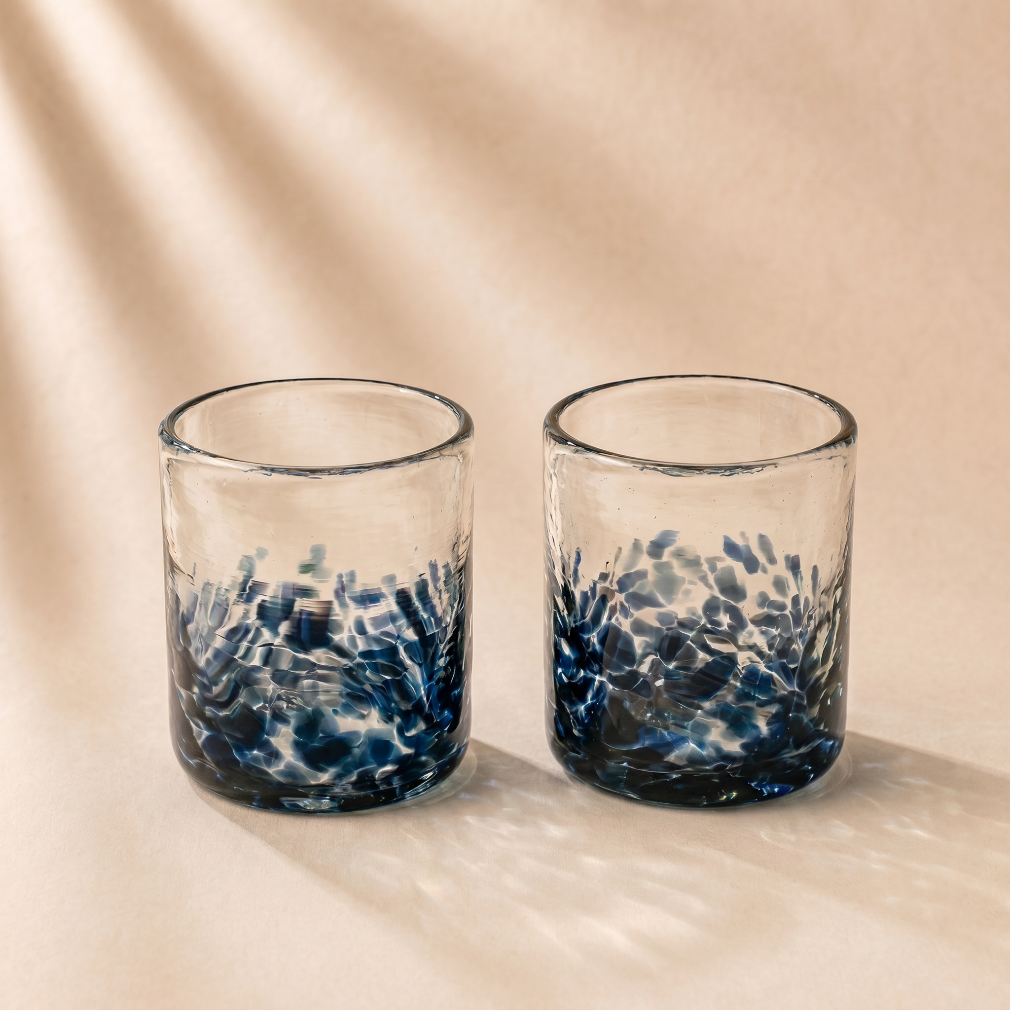 Two clear glass tumblers with a textured, hammered blue pattern concentrated at the bottom, set against a warm beige background with soft leaf shadows.
