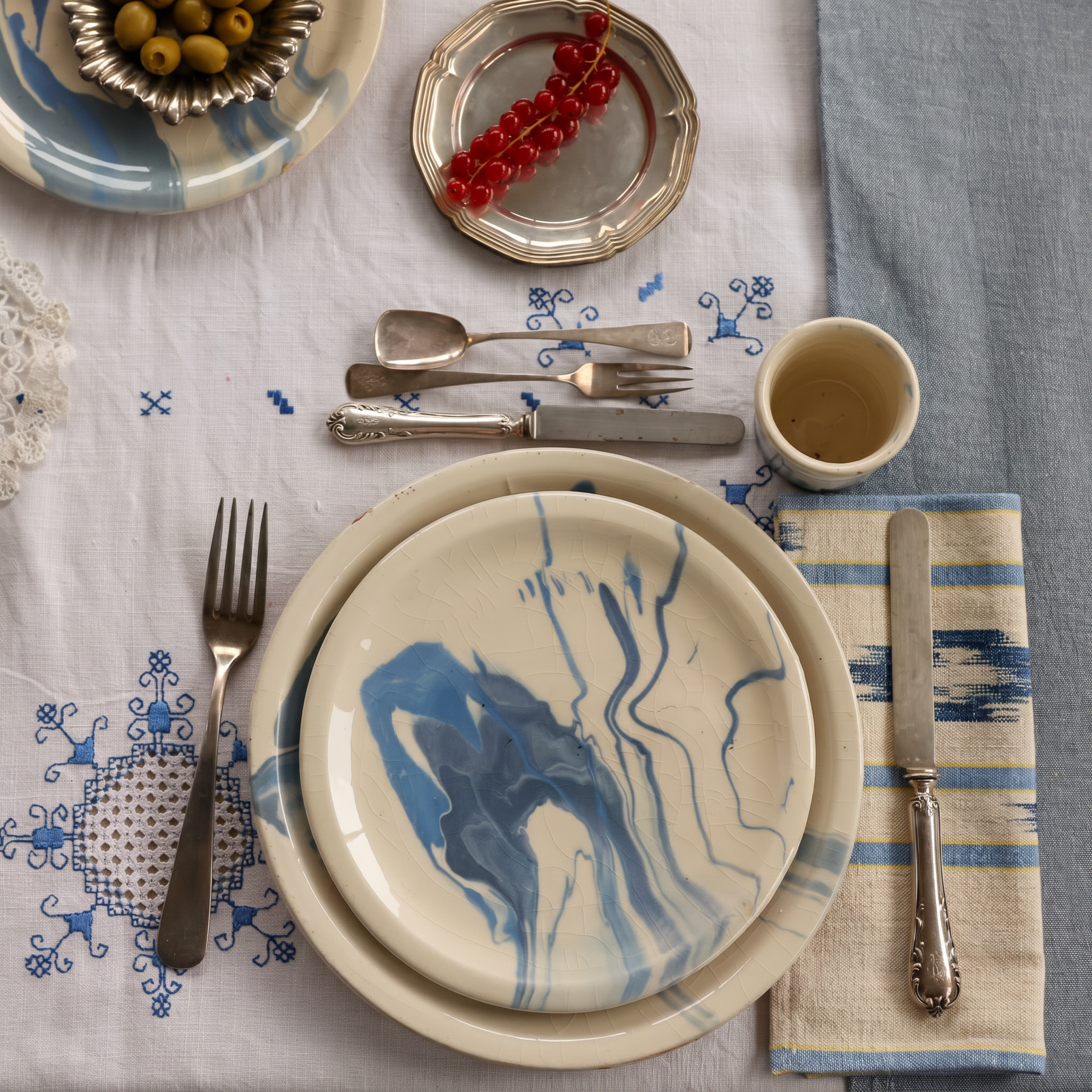 A styled tabletop setting featuring a cream-colored ceramic plate with a blue marbled pattern, layered on a matching plate. The setting includes silver cutlery, a small cream cup, a striped napkin, and a blue-and-white embroidered tablecloth. - lifestyle
