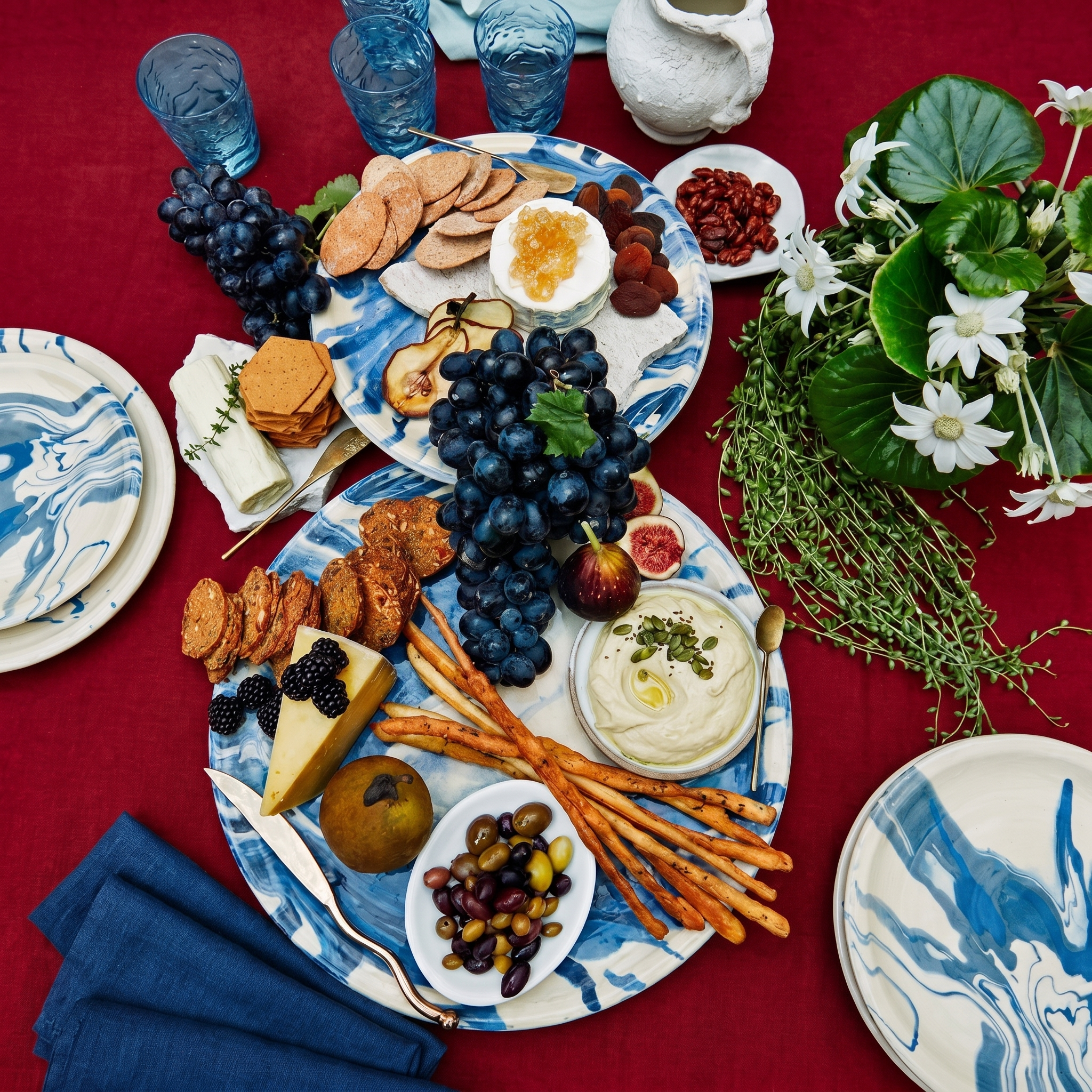 An overhead styled tabletop scene with multiple blue-and-white marbled plates holding charcuterie food, including grapes, crackers, cheese, olives, breadsticks, dips, and small dishes, set on a red tablecloth with blue napkins, blue glassware, flowers, and greenery. - lifestyle
