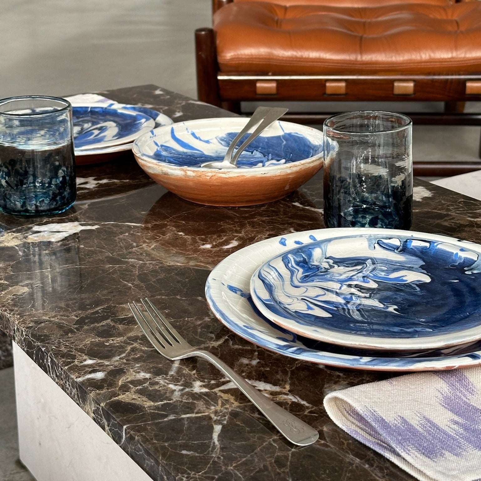 Dining table setting with blue and white dishes, glasses, and cutlery on a marble surface with a brown leather chair in the background.
