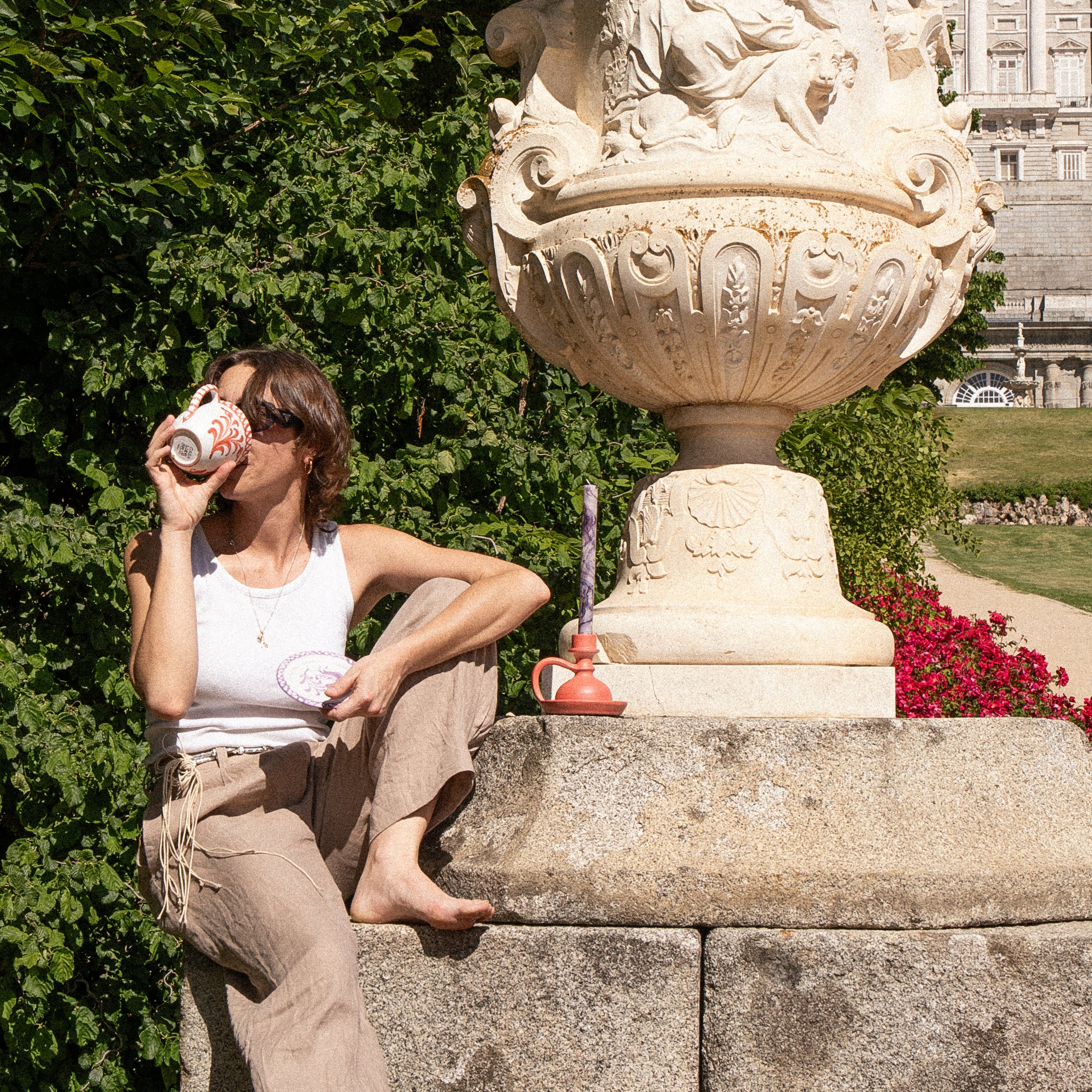Person sitting on a large decorative stone urn in a garden with a building in the background