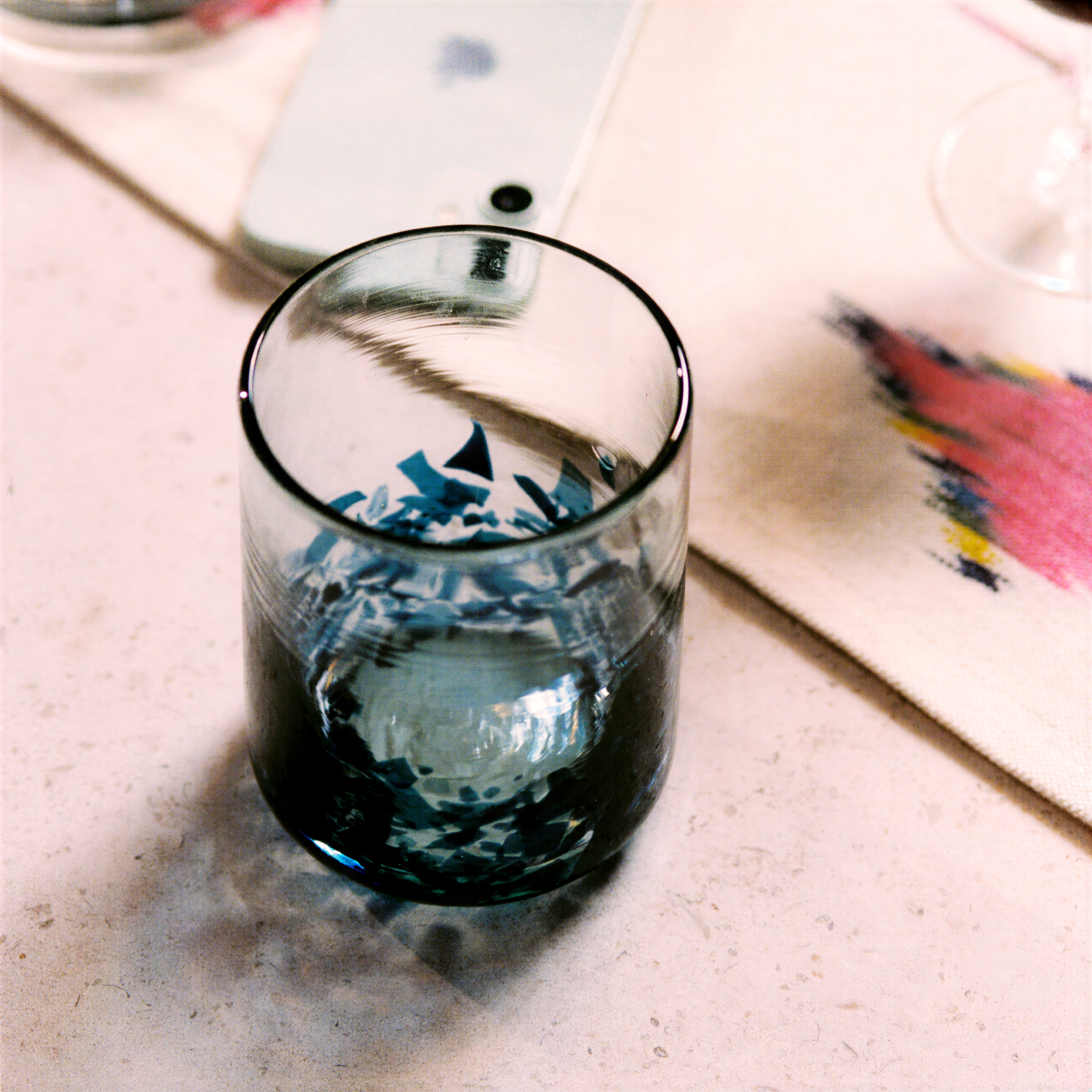 A clear glass tumbler with a dark blue and black abstract pattern near the bottom, photographed on a light tabletop with a phone and placemat in the background. - lifestyle